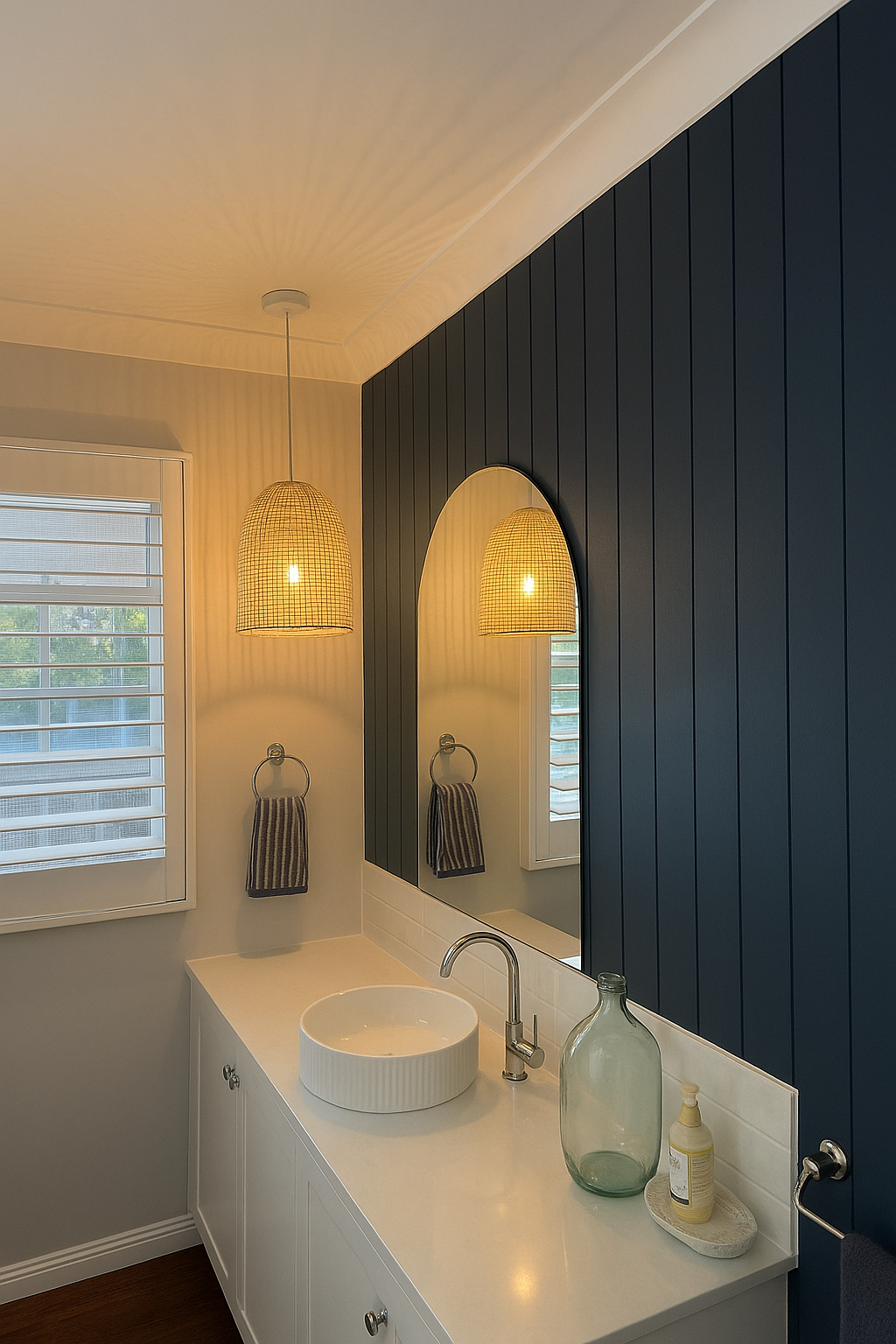 Bathroom vanity area with a white countertop, circular white sink, silver faucet, large mirror, bamboo pendant lights, dark blue vertical wall paneling, window with white blinds, and decorative items including a large glass bottle and soap dispenser.