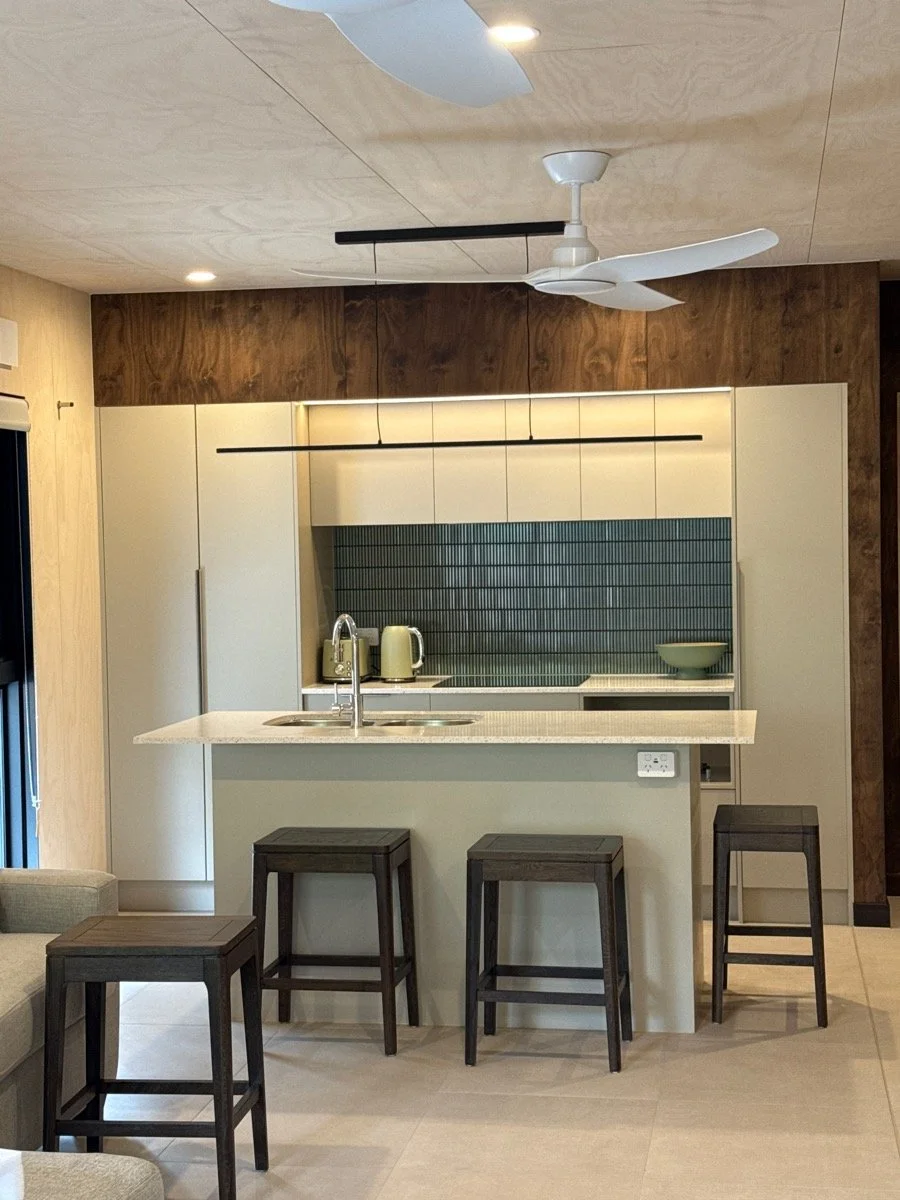 Modern kitchen with white cabinets, dark wood accents, a white countertop island, black barstools, and a ceiling fan.