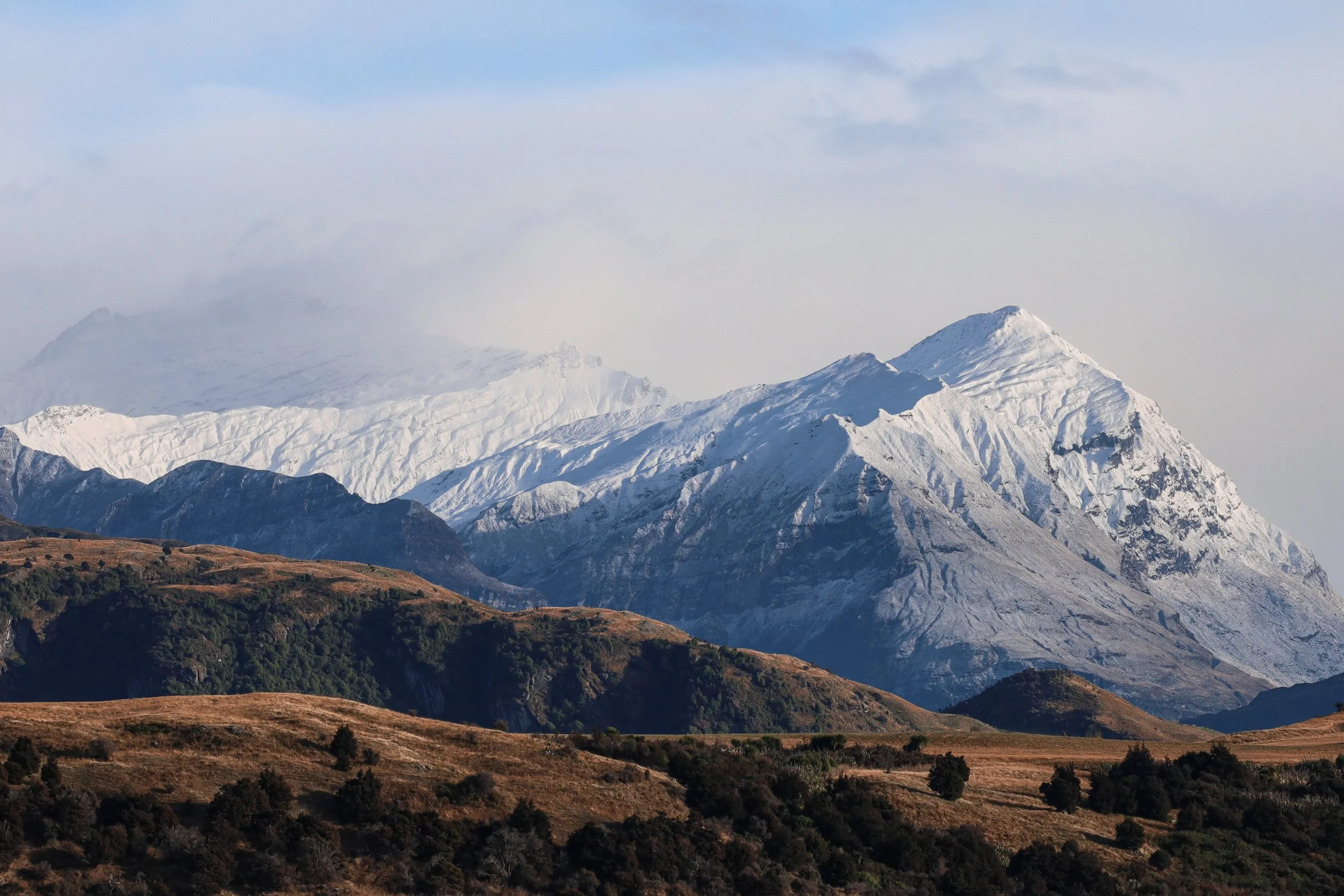 Snow-covered mountains with rolling hills and green vegetation in the foreground. wanaka