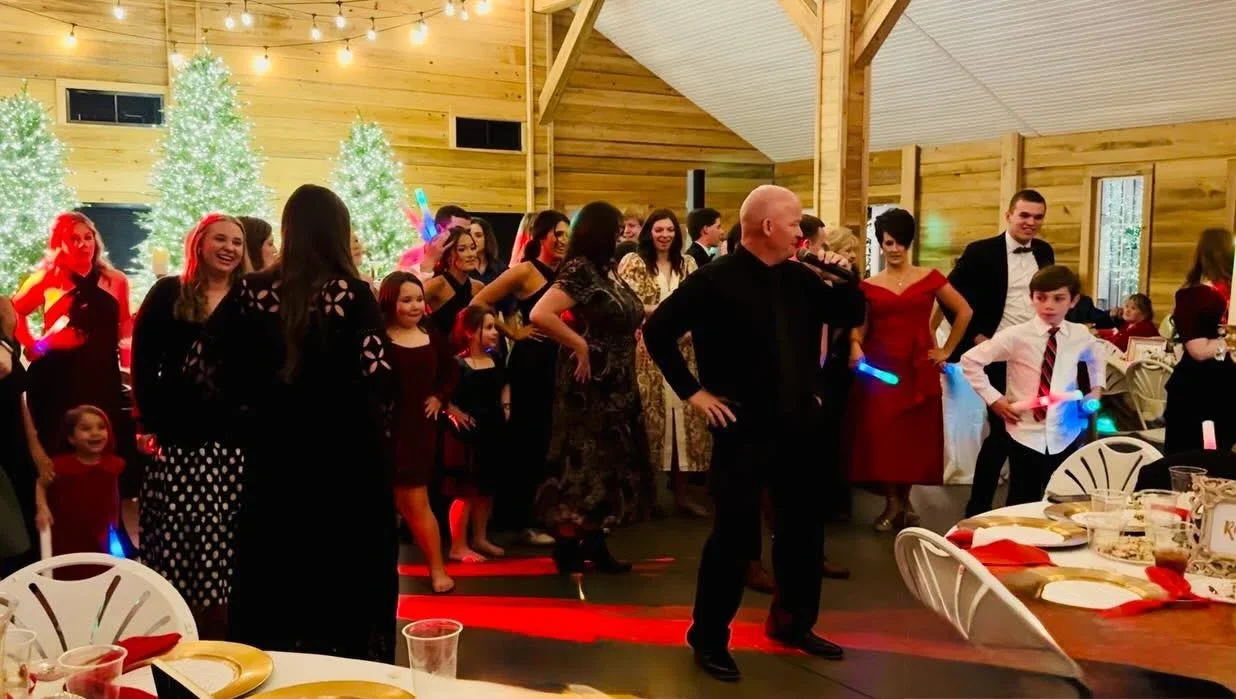 People dancing and celebrating at a holiday party in a decorated wooden hall with Christmas trees, lights, and tables set for a meal.