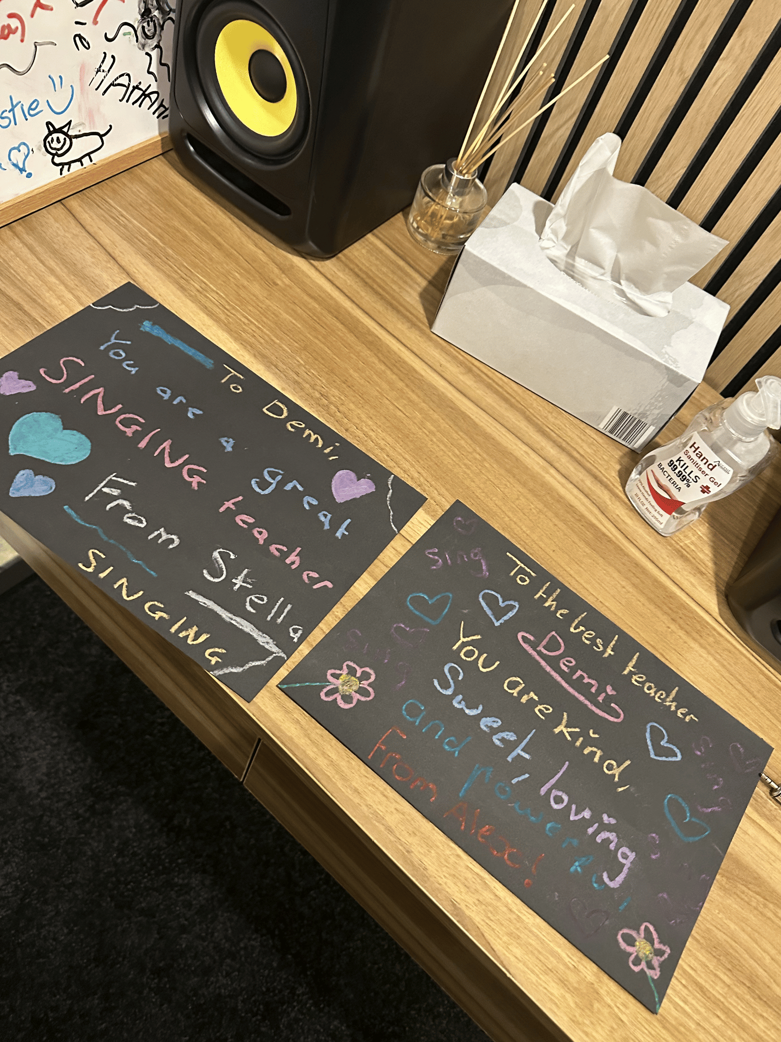 Colorful handwritten notes and drawings on black paper reveal messages about singing and teamwork. Items on the wooden table include a speaker, a tissue box, a bottle of hand sanitizer, and a glass container with sticks.