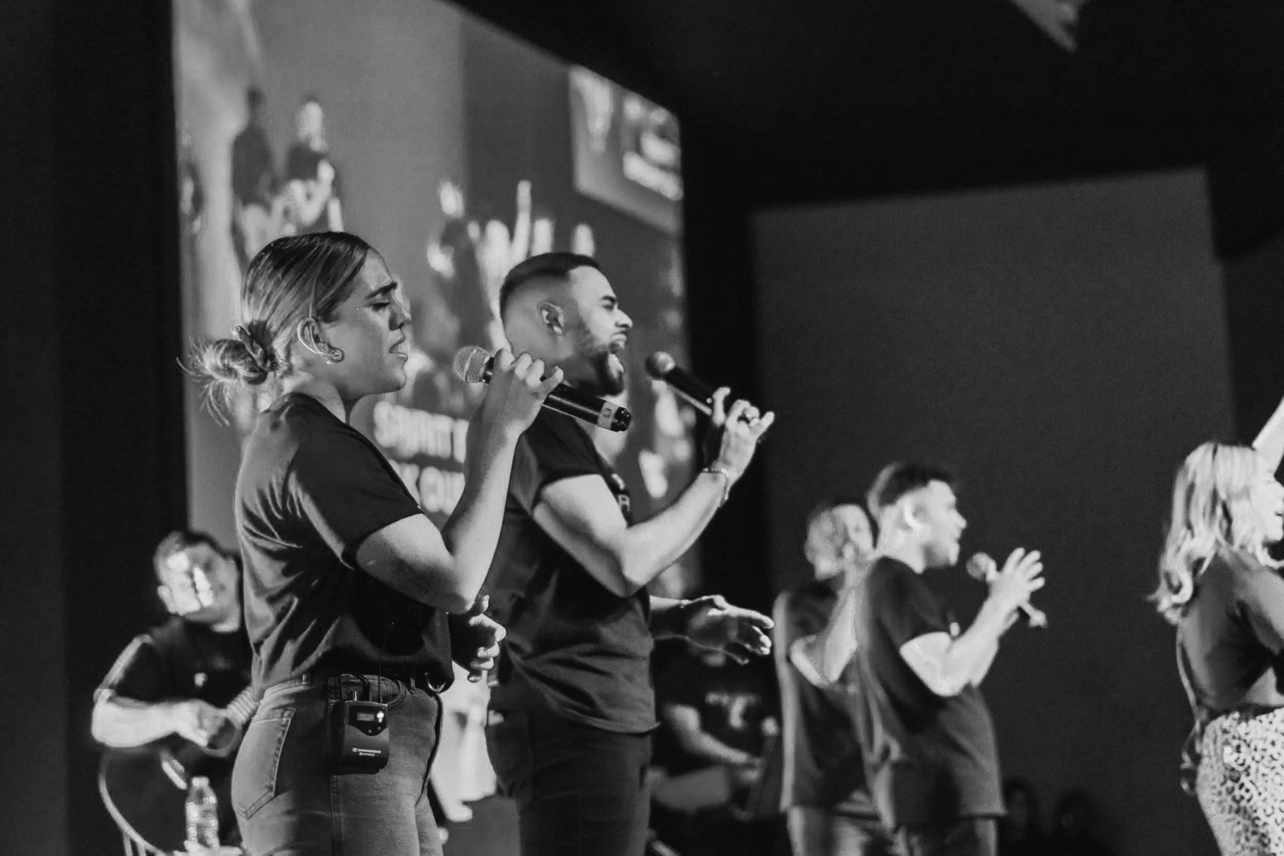 Black and white photo of multiple people singing and playing instruments on stage, holding microphones, with a large screen in the background.