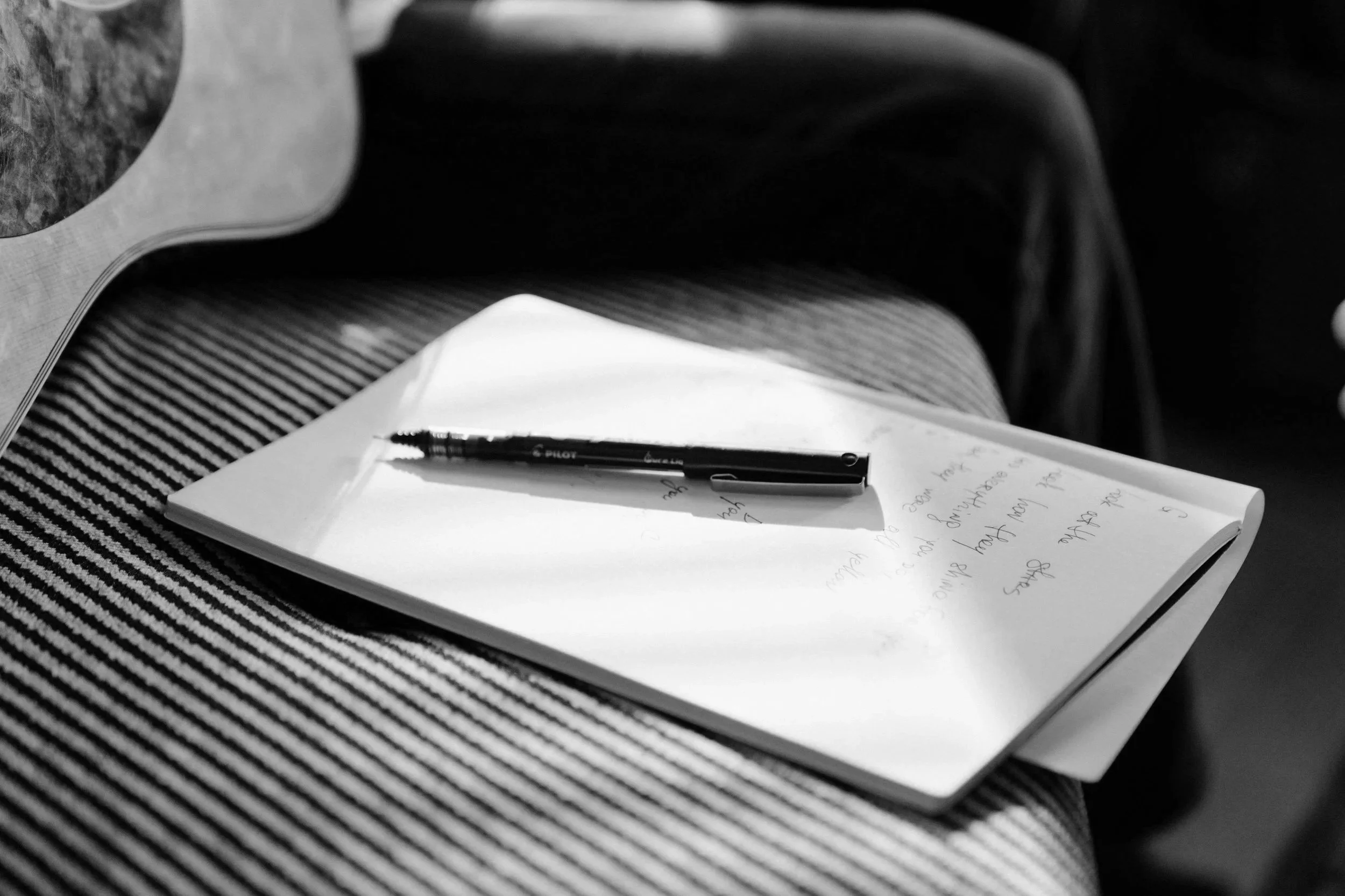 A black ballpoint pen resting on an open notebook with handwritten notes to create a song, placed on a striped fabric surface. 