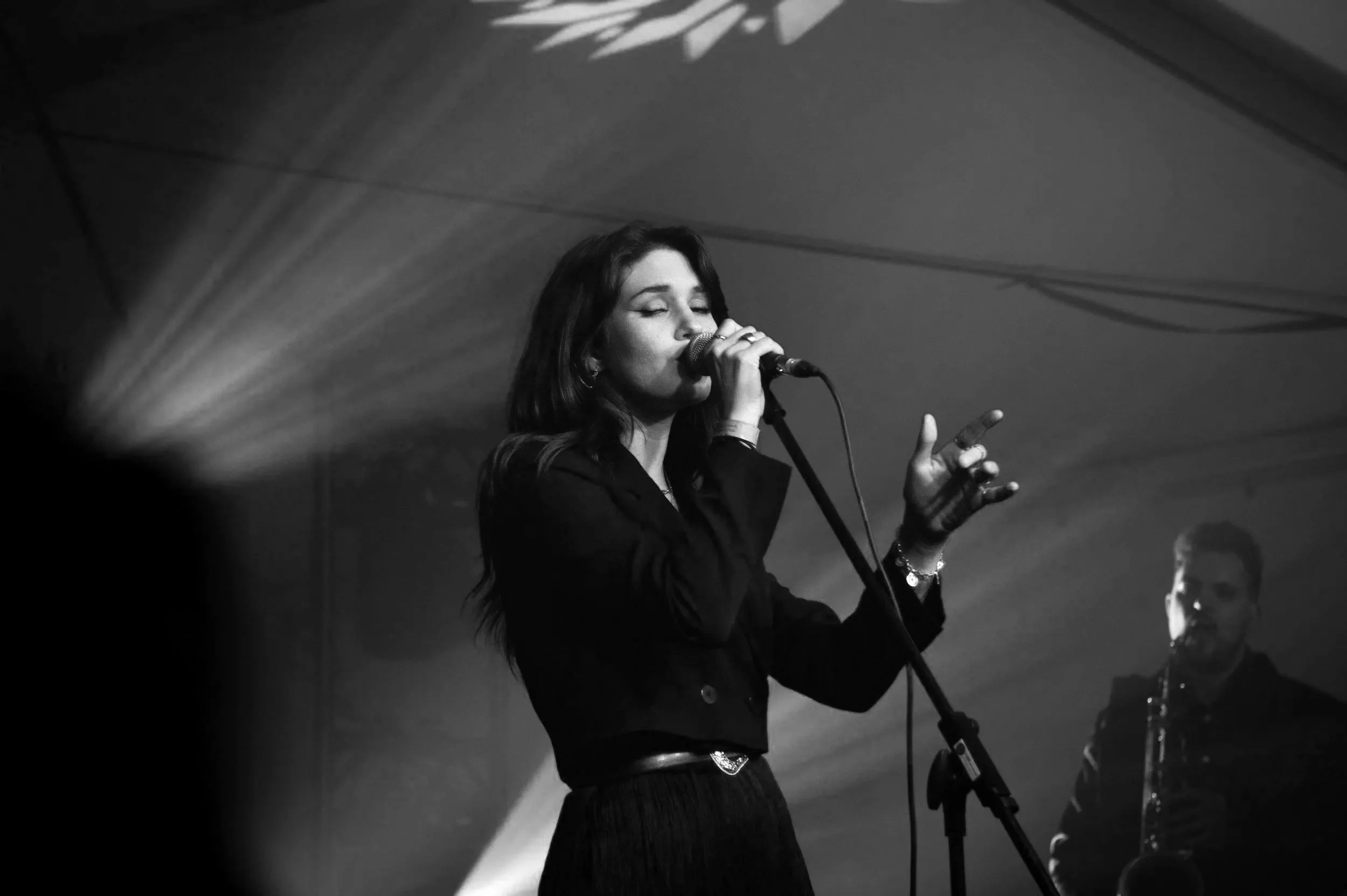 A woman with dark hair singing into a microphone on stage, with a man in the background playing a saxophone.