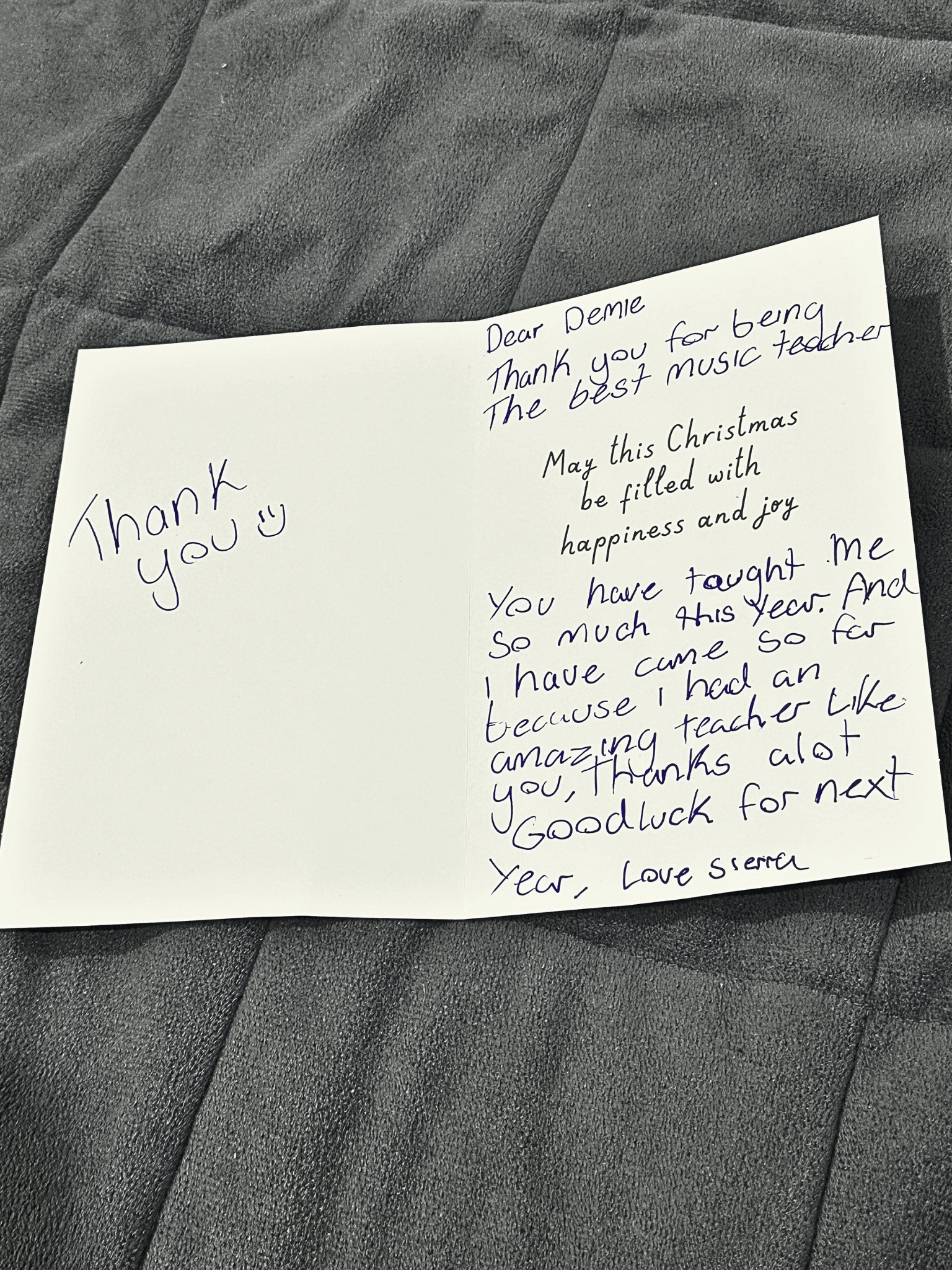A handwritten note on white paper placed on a black leather surface. The note contains a thank you message to Demie for being a music teacher and wishes for a joyful Christmas and good luck in the new year.