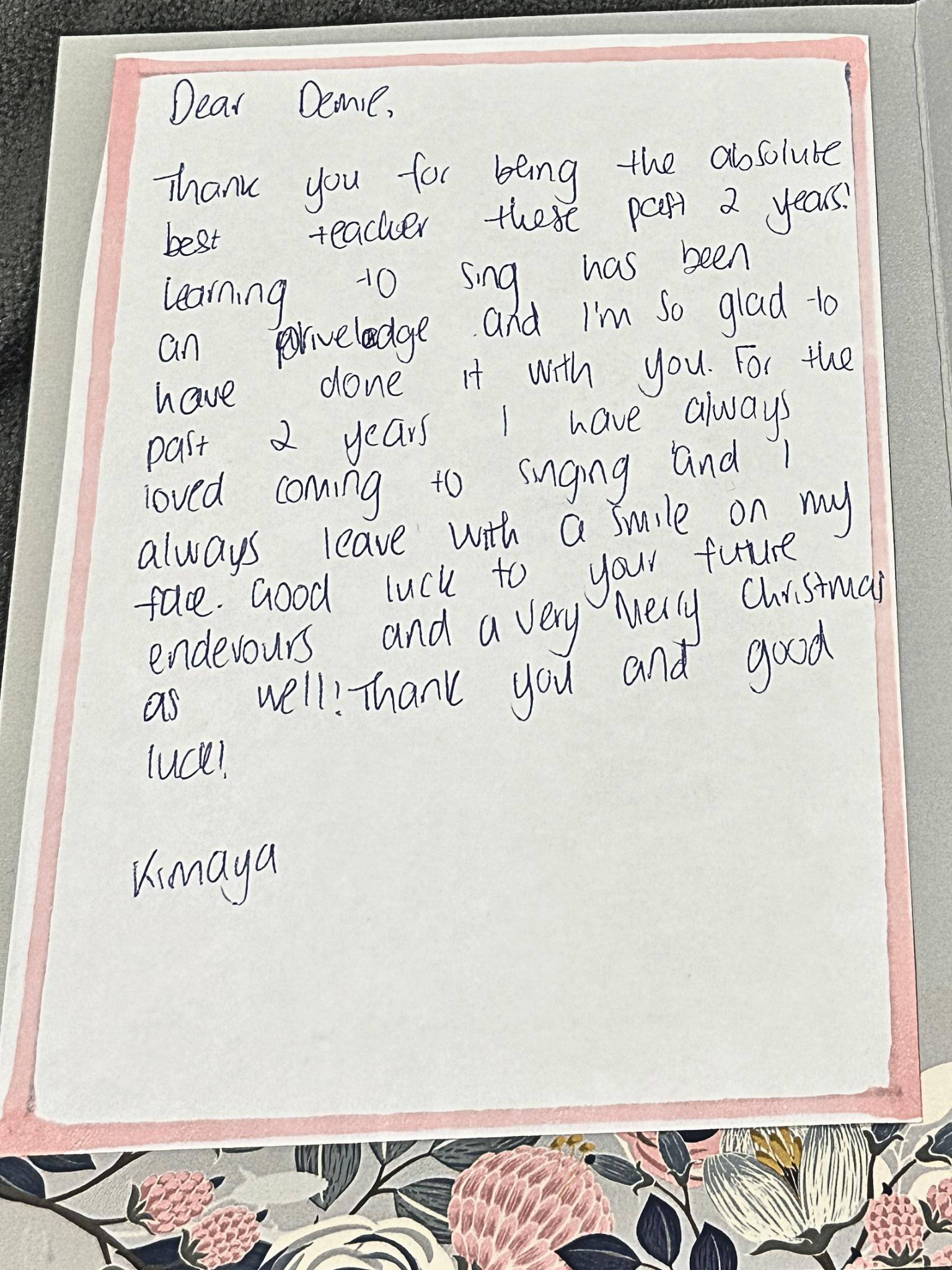 A handwritten letter on white paper with pink borders, placed on top of a floral-patterned surface. The letter is addressed to Danielle and expresses gratitude for being the best teacher and for singing lessons.