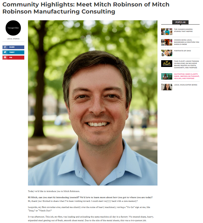 Photograph of a smiling man with short brown hair, wearing a light blue shirt, standing outdoors in a park with green trees in the background.