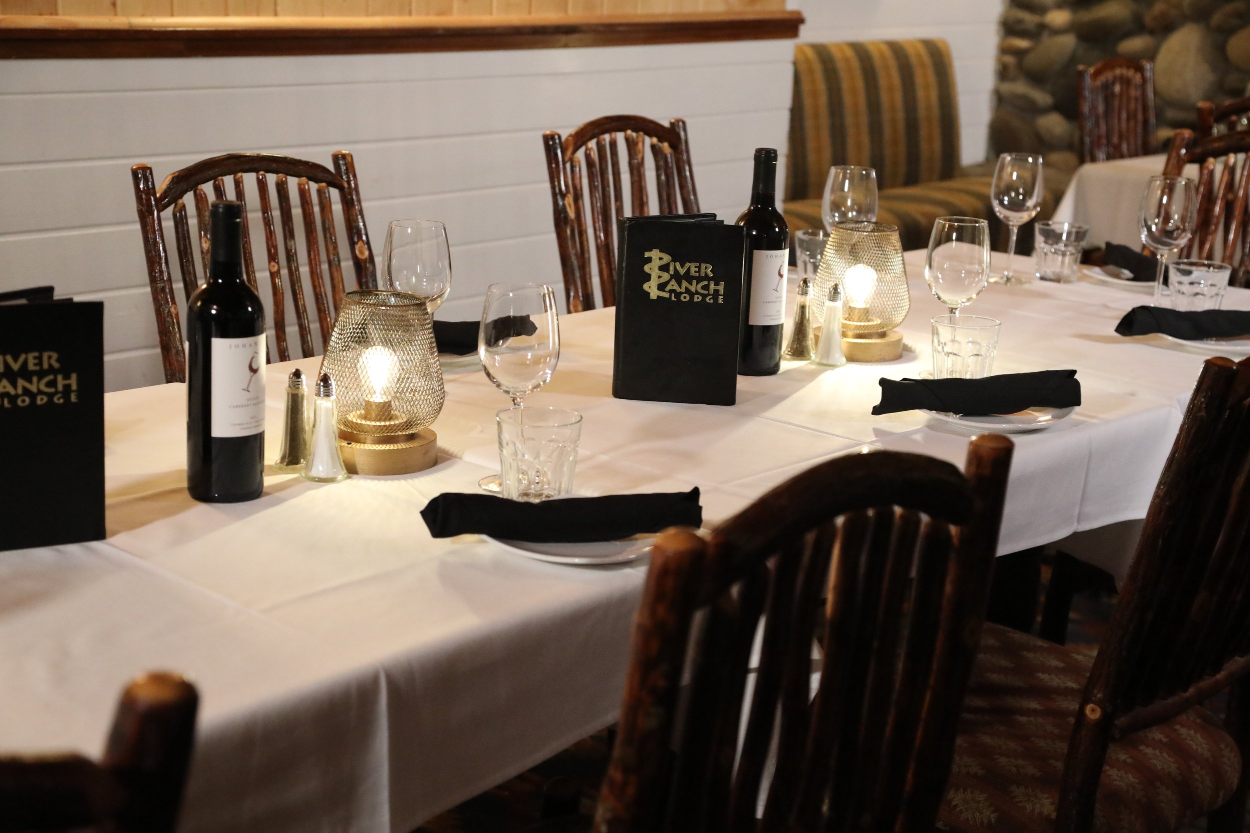 A dining table set for a meal with wine bottles, glasses, black napkins, and decorative lamps in a cozy restaurant setting.