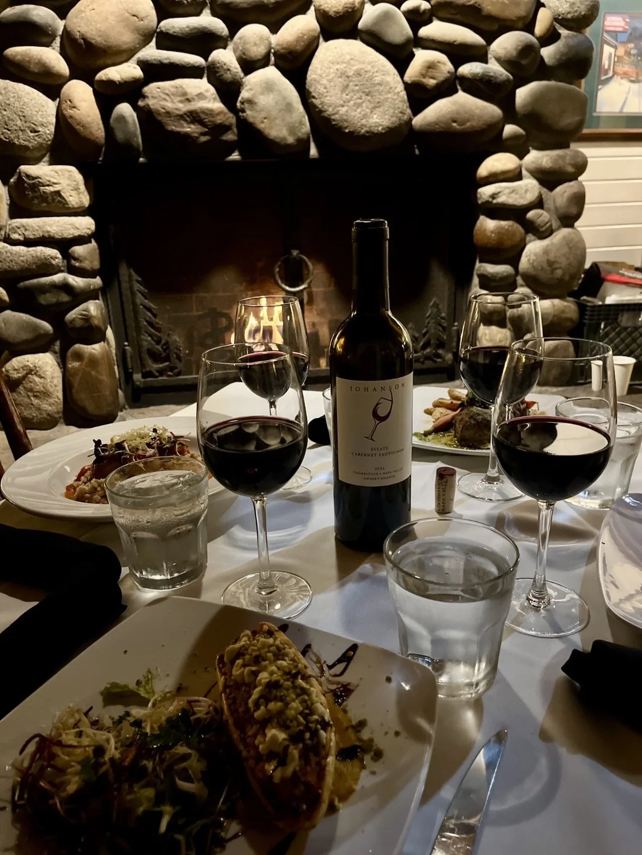 A table set for dinner with a fireplace in the background. The table has a bottle of red wine, six wine glasses filled with red wine, plates of food, and glasses of water.