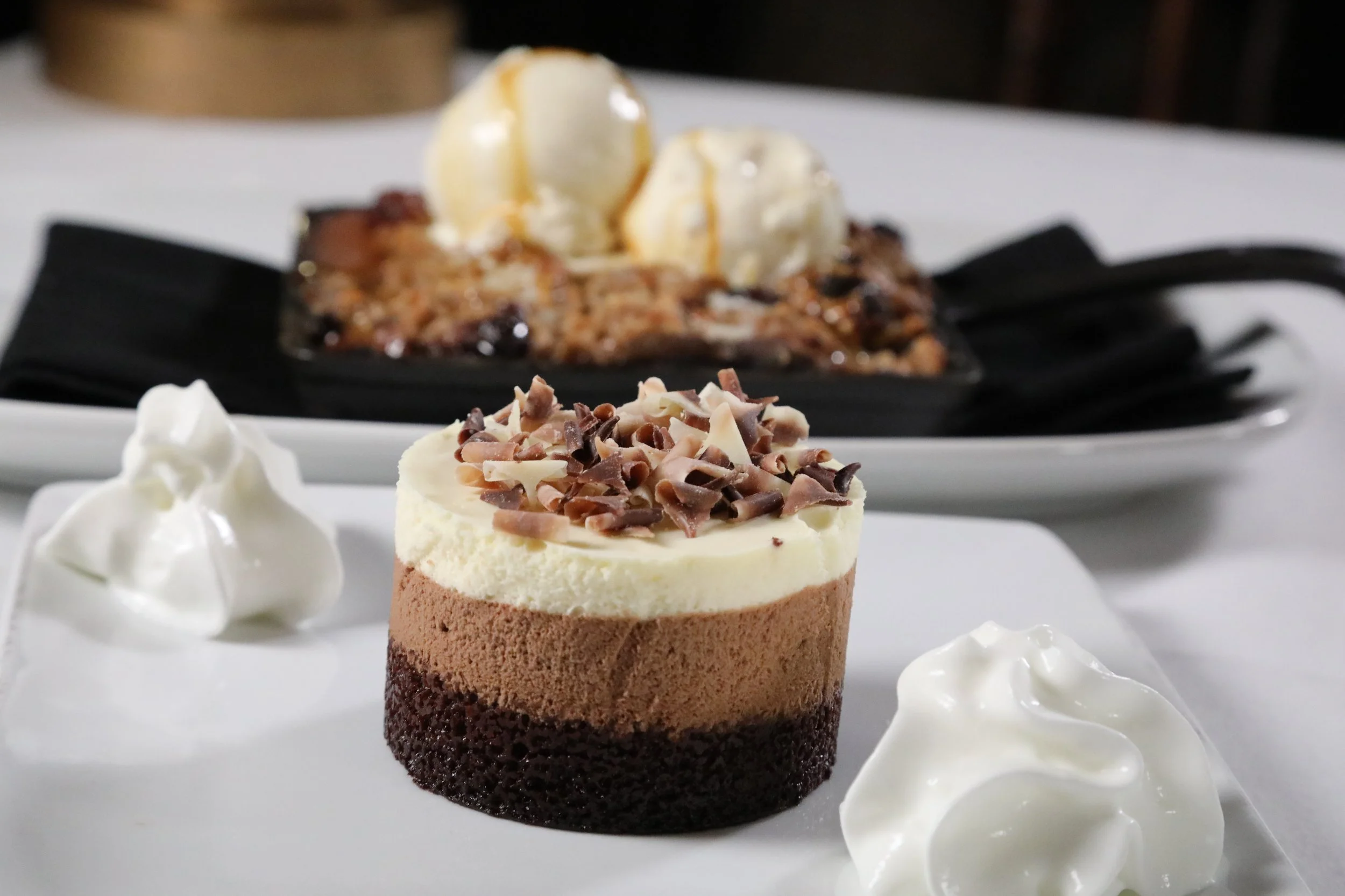 A layered chocolate mousse dessert topped with chopped chocolate shavings, served with dollops of whipped cream on a white plate, with an apple crumble dessert topped with two scoops of vanilla ice cream in the background.