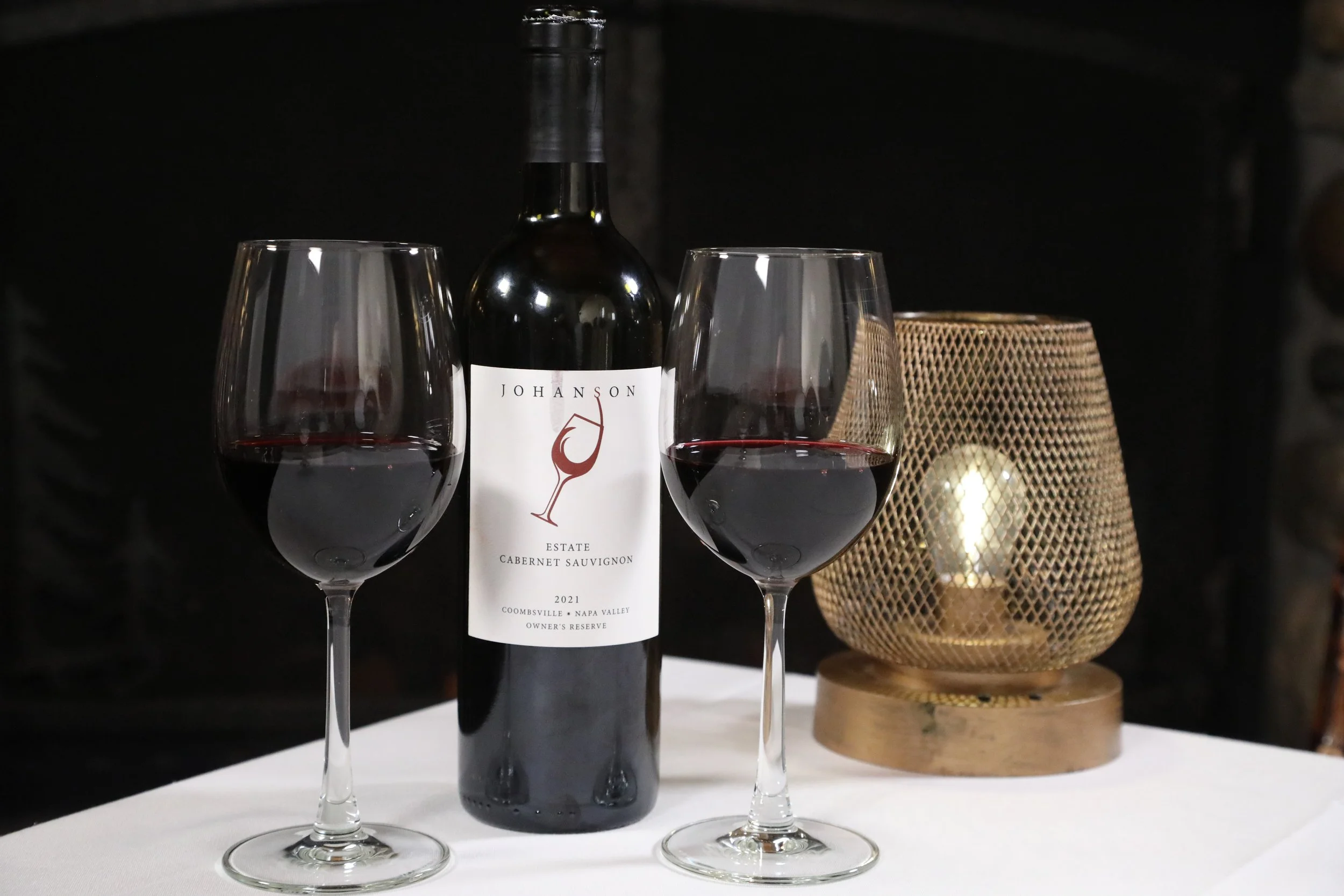 A bottle of Johan Sin Estate Cabernet Sauvignon 2021 wine with two filled wine glasses on a white table, and a candle holder with a lit candle in the background.