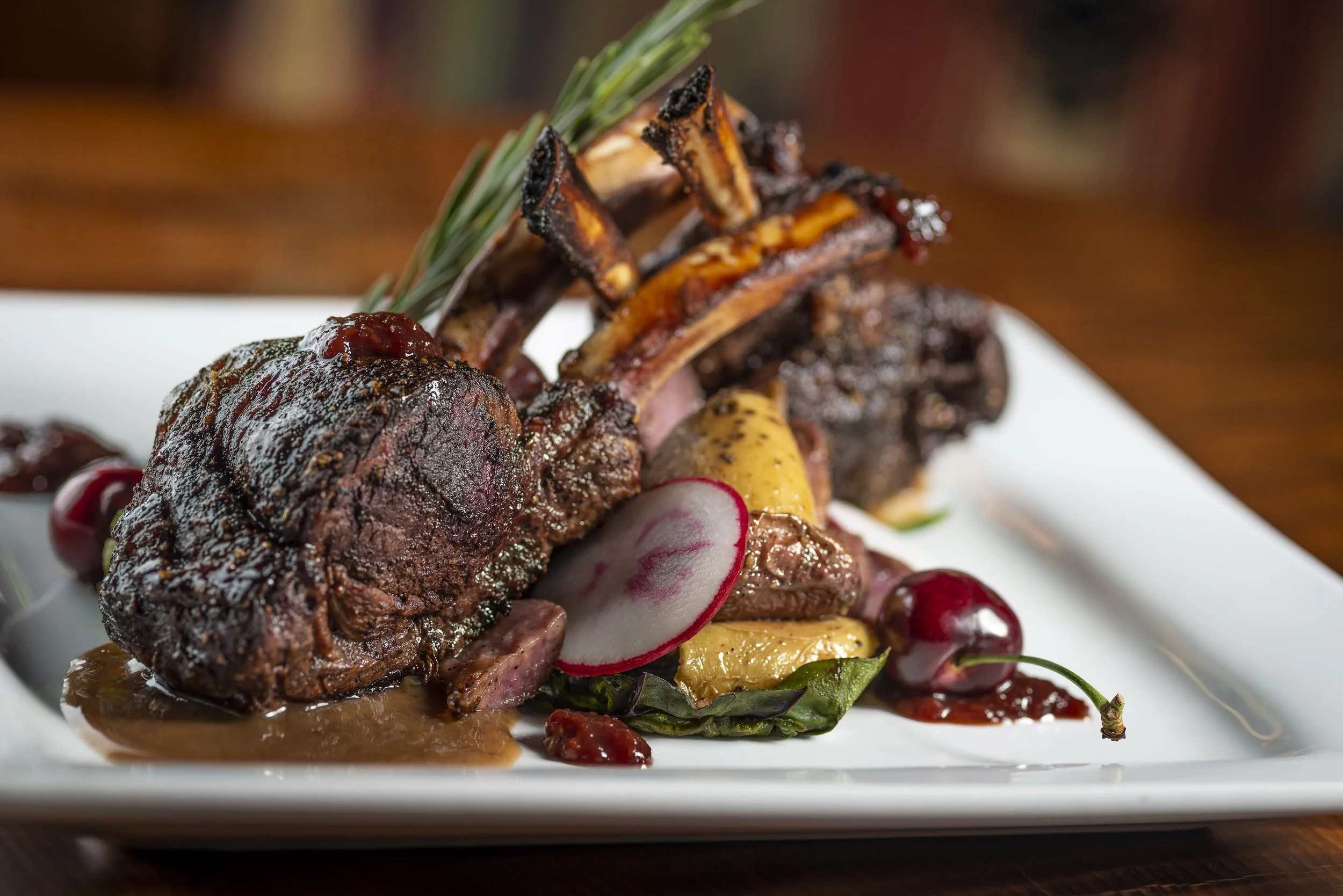 Roasted rack of lamb with herbs, cherry garnish, and roasted vegetables on a white plate.