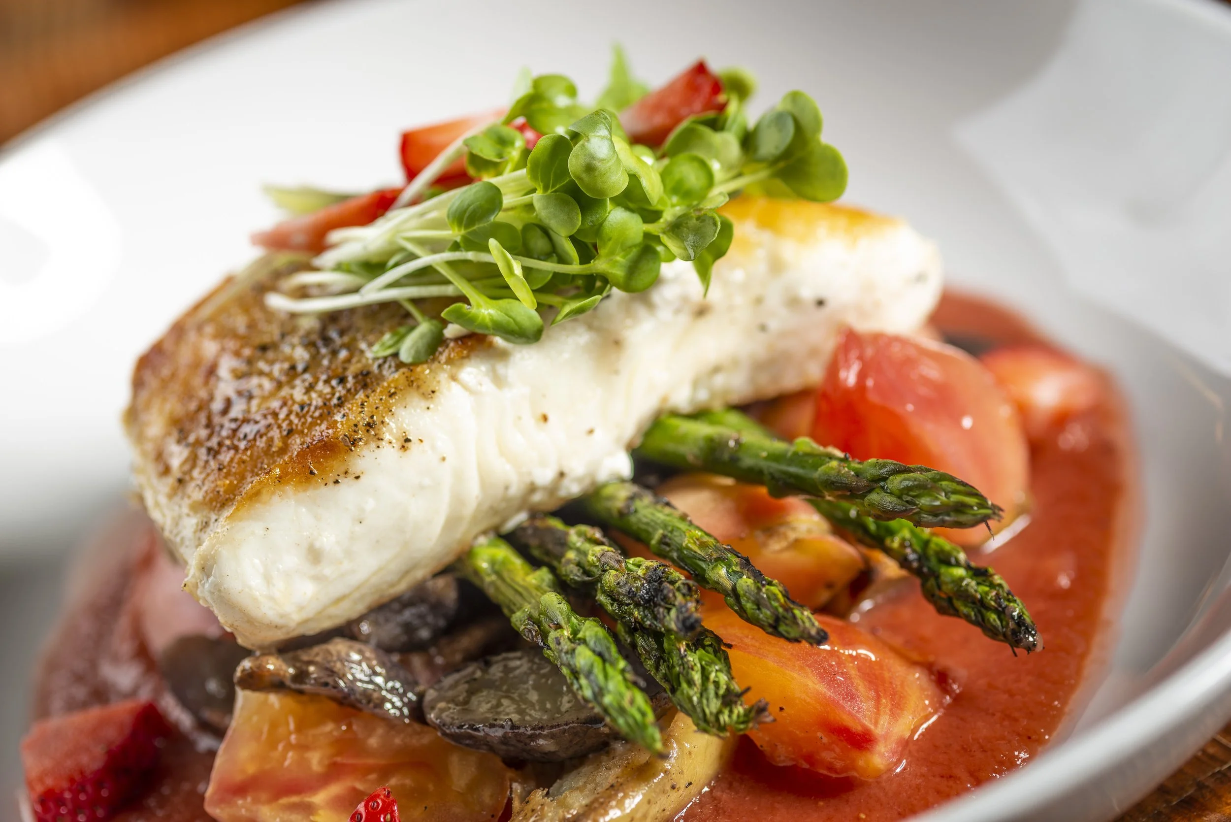 A plate with baked fish fillet, topped with microgreens, served over asparagus, tomatoes, and vegetables in tomato sauce.