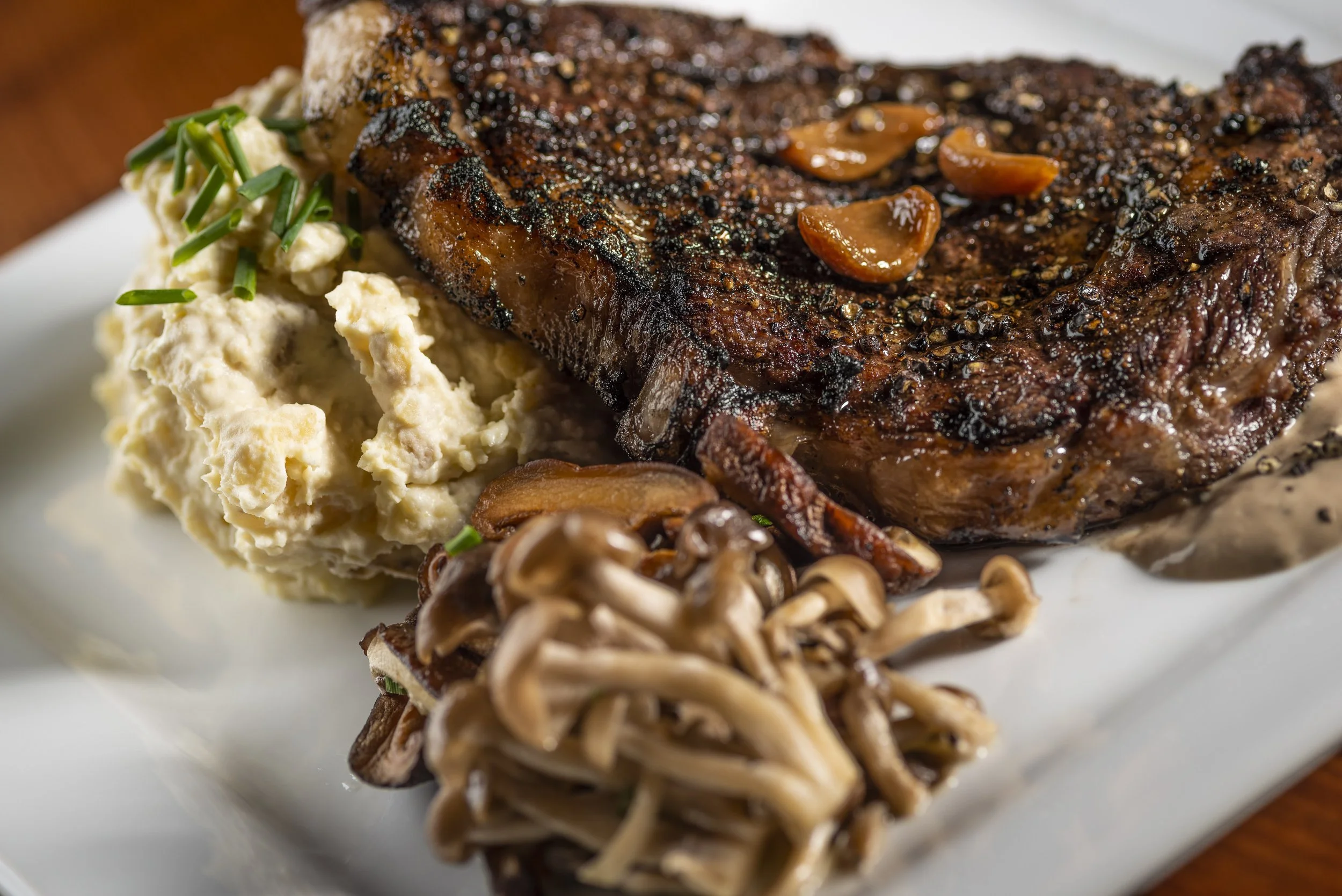 Close-up of a grilled steak with garlic and pepper, alongside mashed potatoes with chives, sautéed mushrooms, and gravy on a white plate.