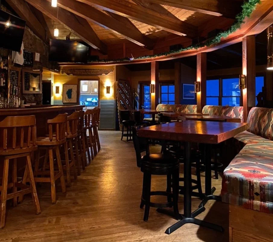 Empty cozy bar and dining area with wooden furniture, a bar counter on the left, a row of chairs, booth seating along the windows, warm lighting, and a decorated ceiling with green garland.