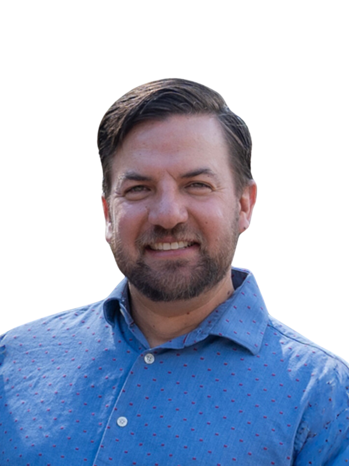 A smiling man with a beard and brown hair, wearing a blue button-up shirt with small red polka dots, standing outdoors.
