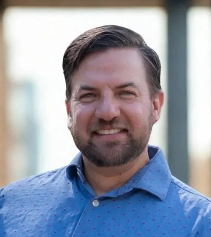 A man with a beard and short brown hair, smiling, wearing a blue collared shirt, outdoors