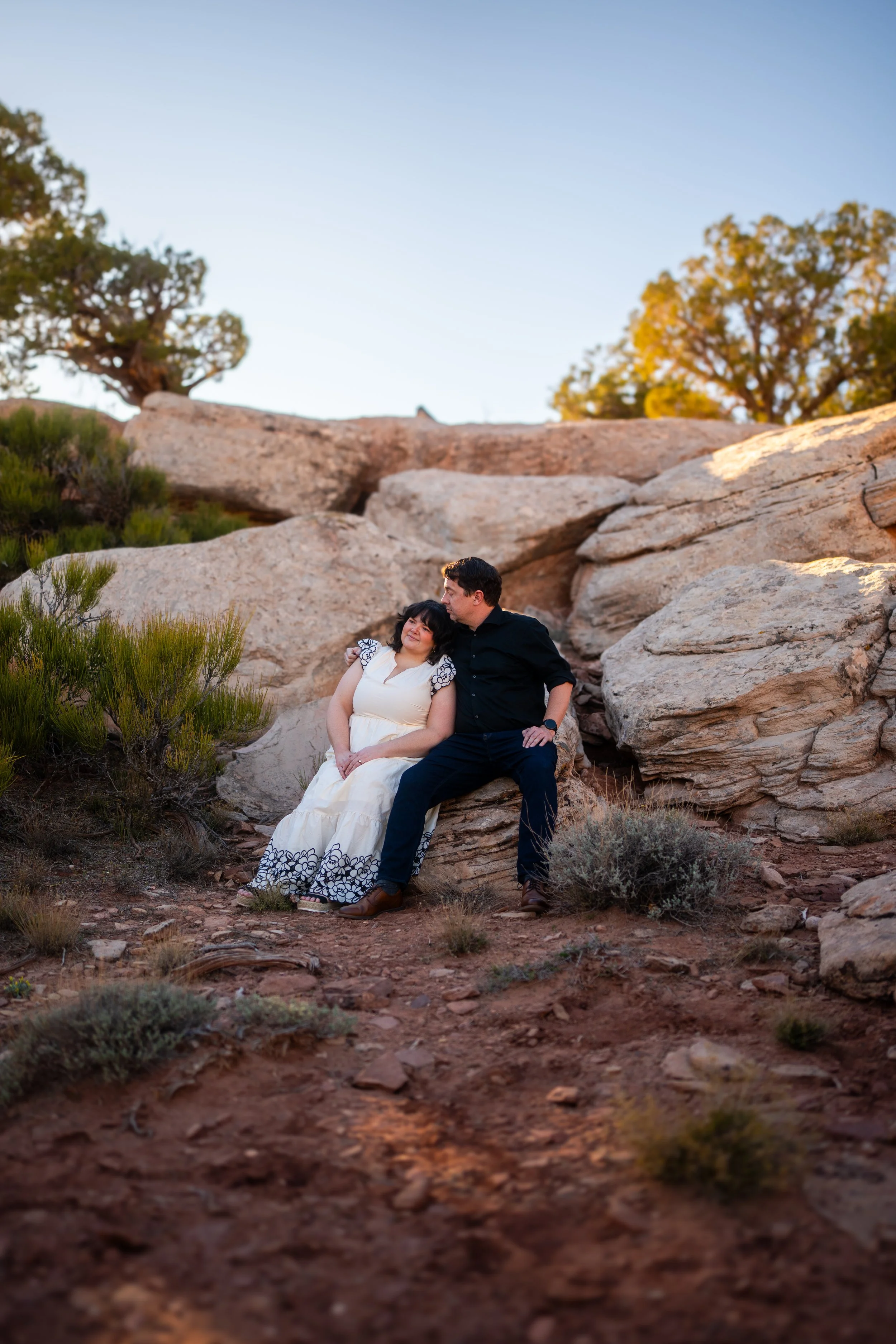 A couple sitting on a large rock in a desert landscape with shrubs and trees, during sunset.