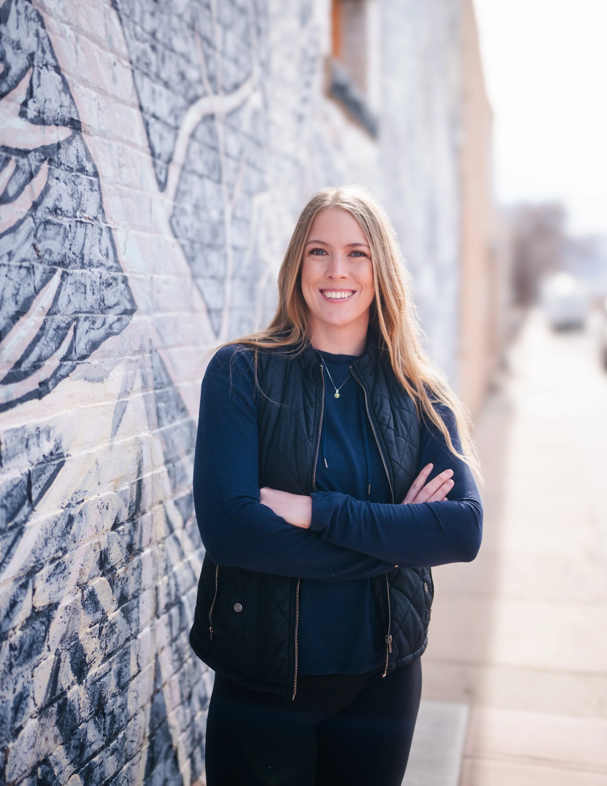 A woman with long blonde hair smiling and standing confidently with her arms crossed, leaning against a brick wall with mural artwork, in an outdoor urban setting.