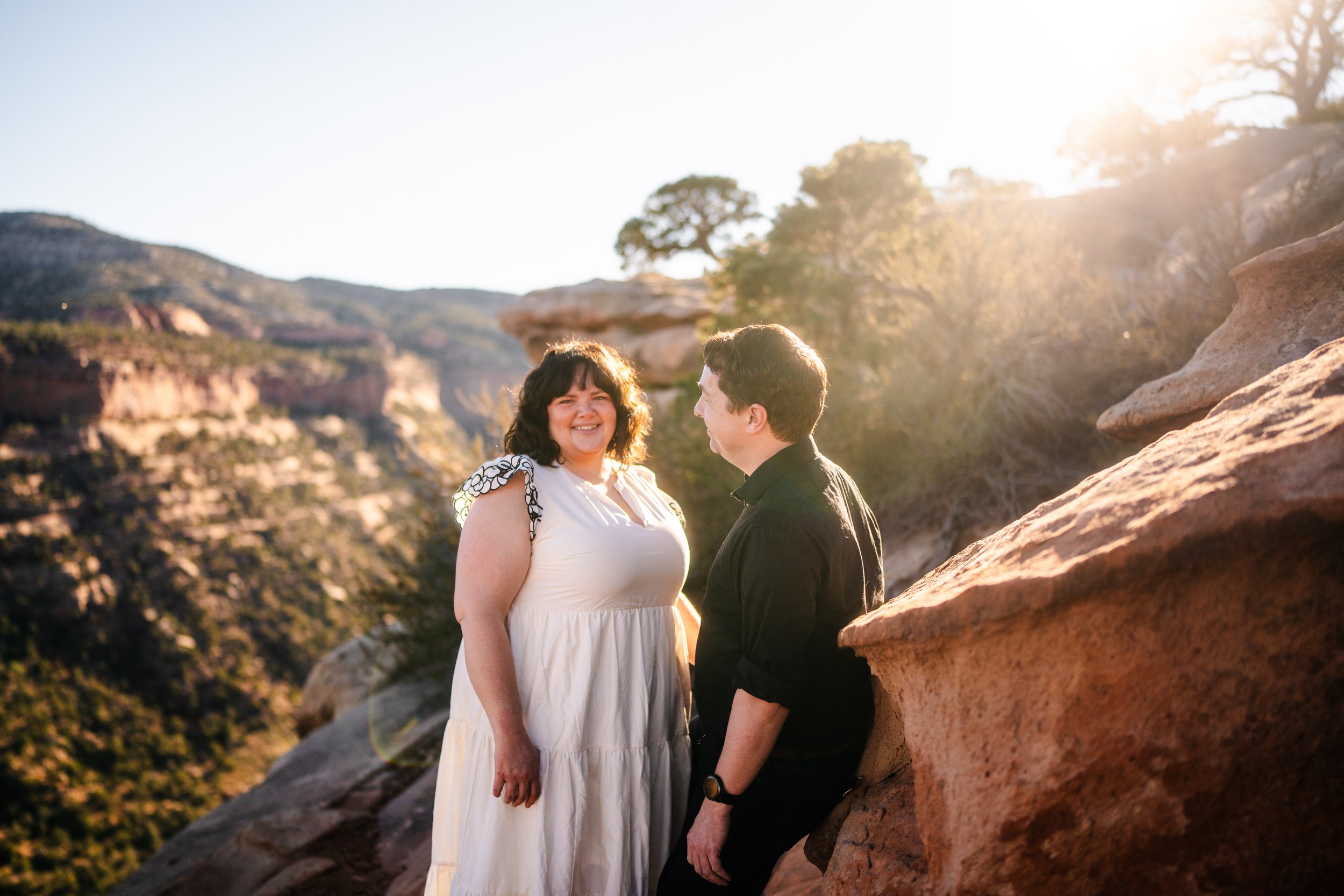 A couple standing on a rocky ledge outdoors during sunset, smiling and looking at each other, with a mountainous landscape in the background.