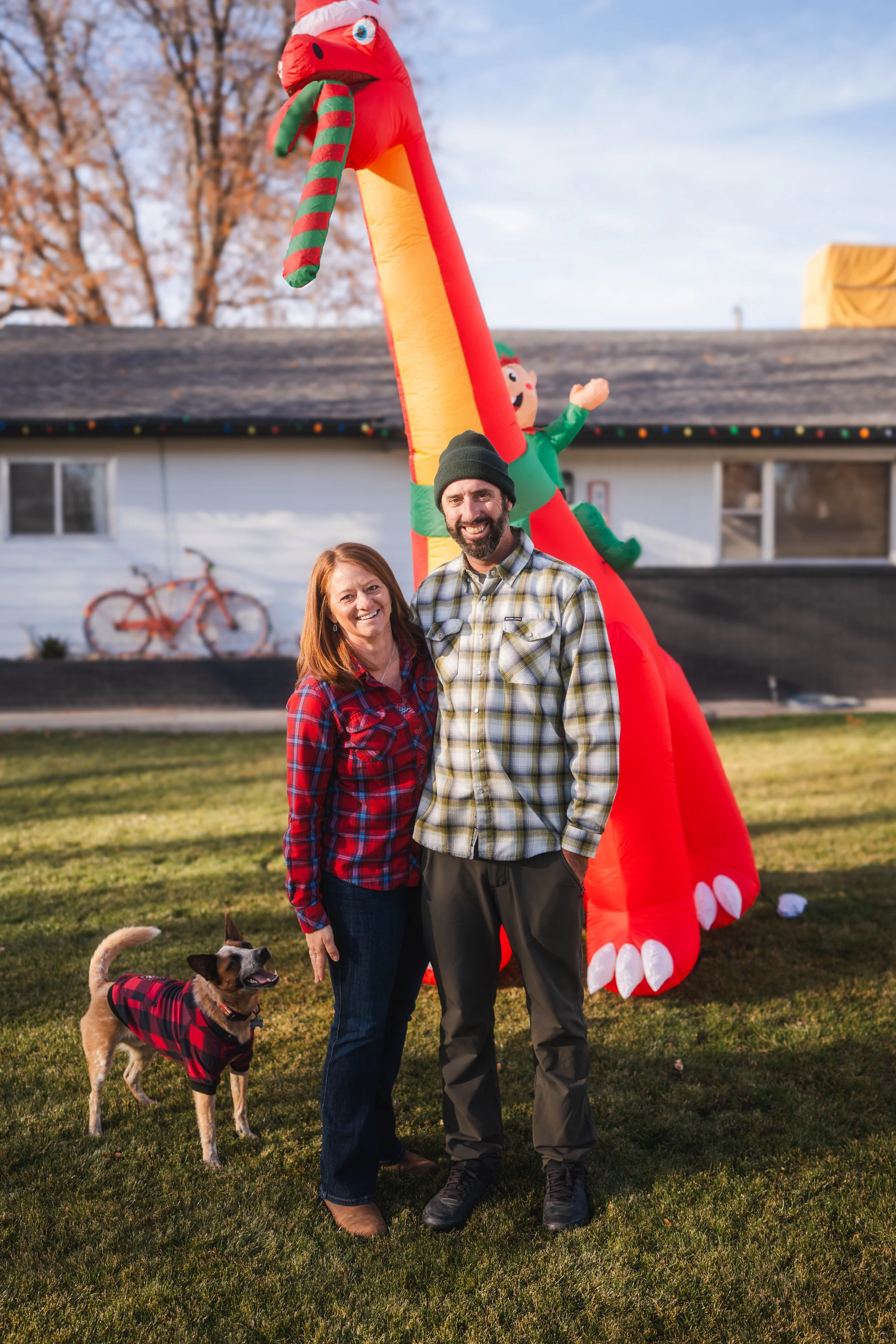 A smiling couple and their dog in front of a large inflatable Christmas-themed decoration, including a red and yellow zipper with a green elf on top, outdoors on a grassy lawn.