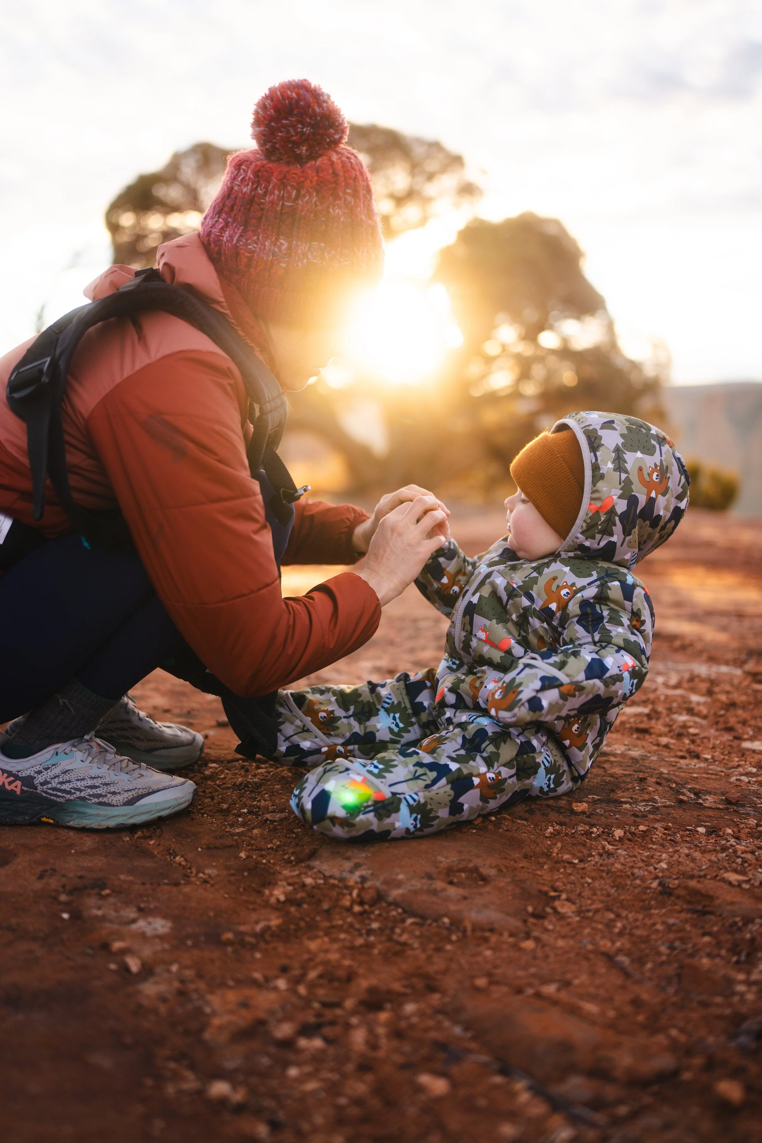 A woman and a young child outdoors at sunset, the woman is kneeling and holding the child's hand, both are dressed warmly in jackets and hats, with the sun setting behind trees in the background.