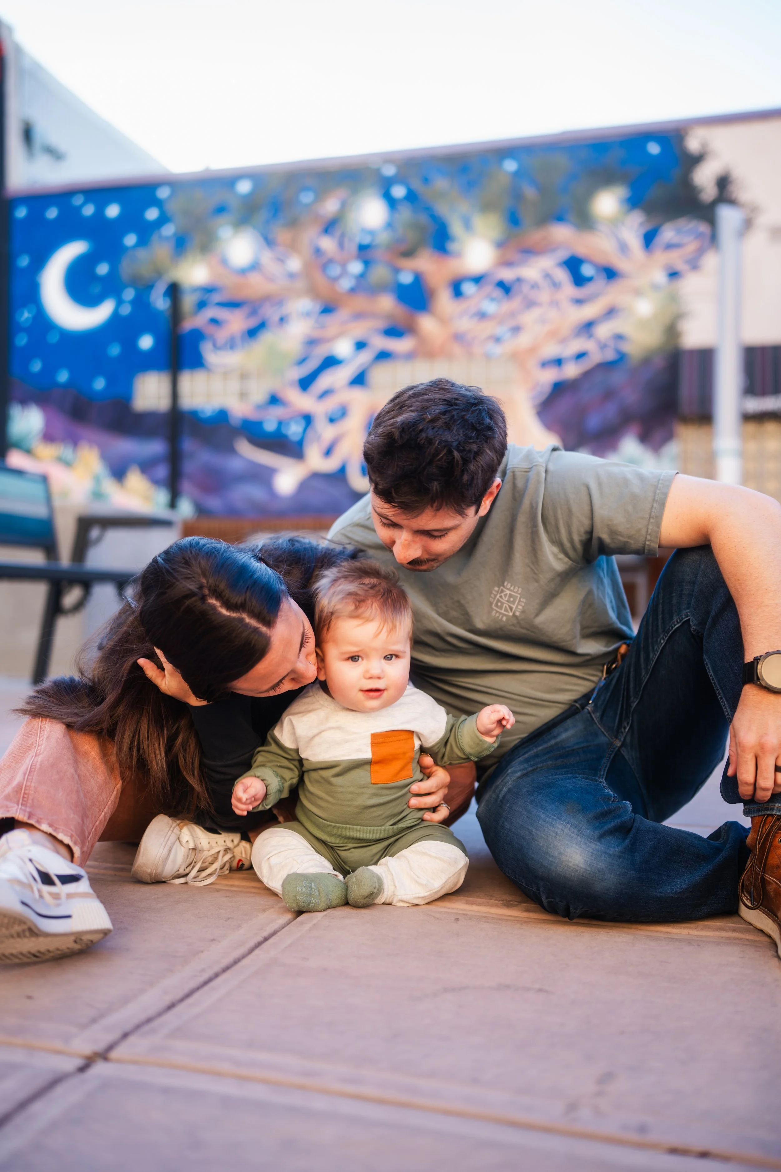 A family of three, including a woman, man, and toddler, sitting on a wooden deck outdoors, with a colorful mural of a tree and night sky in the background.