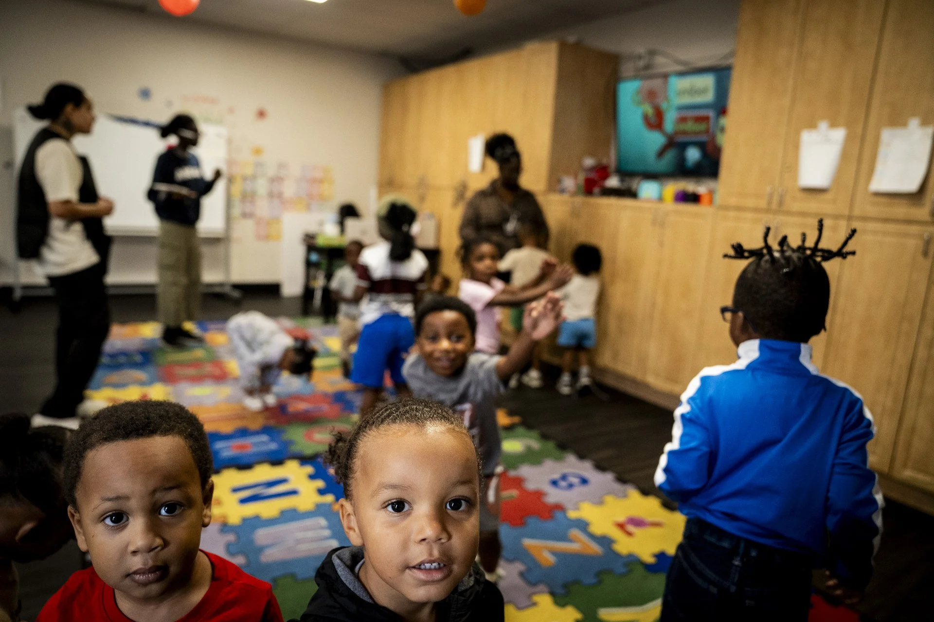 Children in a classroom, some interacting with each other, with teachers and educational materials present.