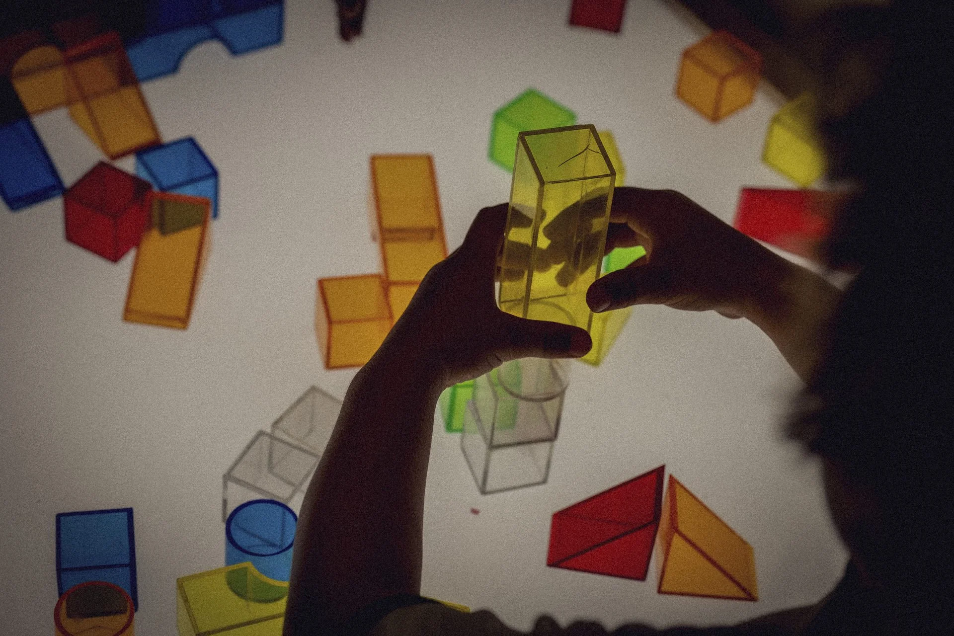 A child playing with translucent, colorful geometric blocks on a lightbox surface, creating shadows and cast reflections.
