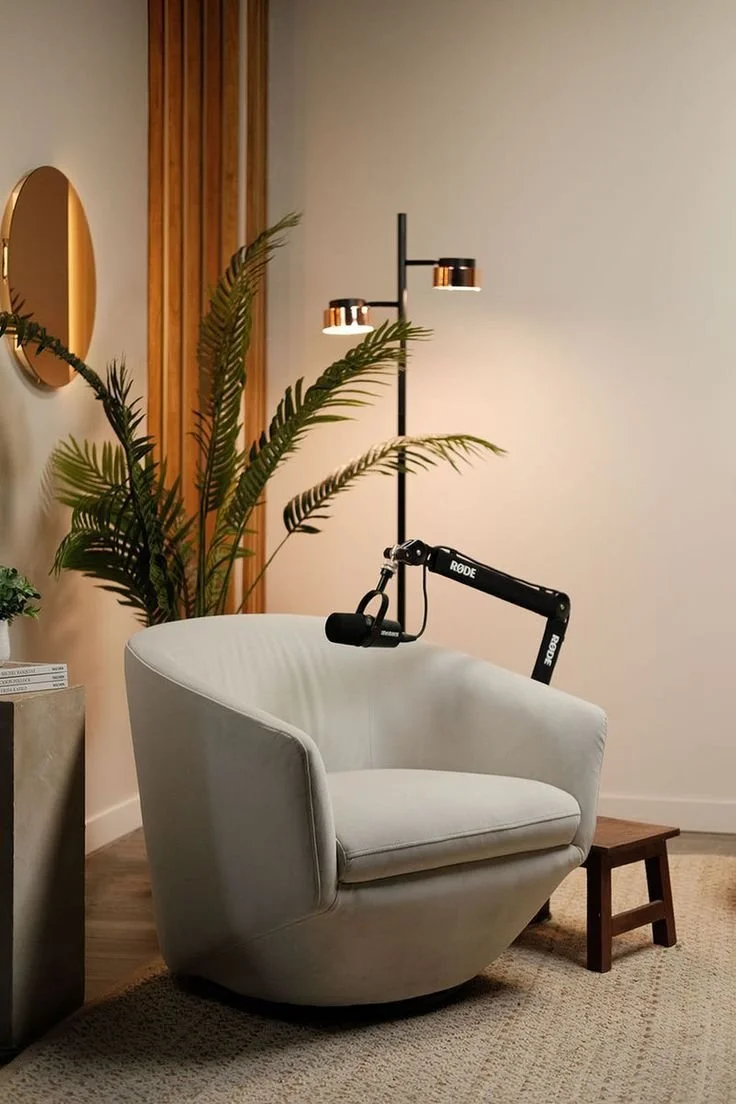 A cozy armchair with a microphone attachment and a small wooden stool beside it in a modern living room. There is a tall floor lamp, a large plant, and a mirror on the wall.