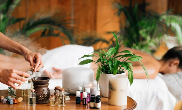 A person lying on a massage table in a spa, receiving a massage with their face turned to the side. The scene includes aromatherapy oils, candles, and potted plants on a wooden table in the foreground.
