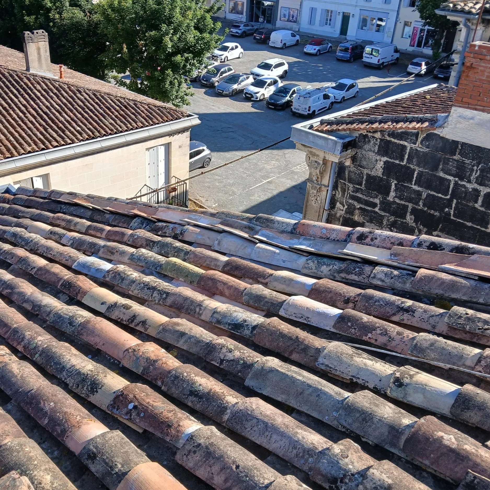 Vue du toit en tuiles anciennes avec de la mousse, regardant vers une cour avec un parking rempli de voitures et au-delà, une rue avec plus de voitures et des bâtiments.