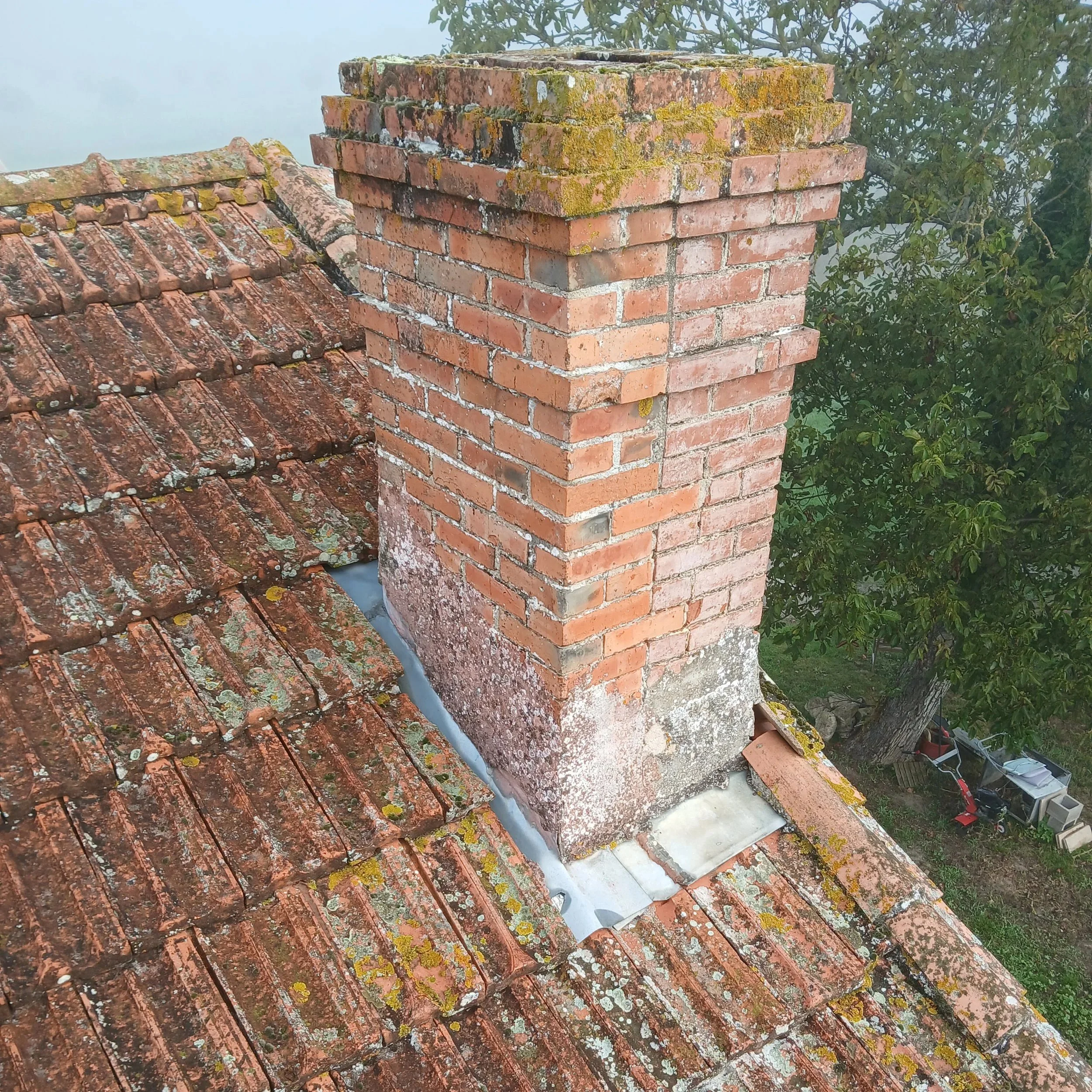 Cheminée en briques sur un toit en tuiles, avec mousse et lichens, vue du haut, entourée d'arbres et de matériel de jardinage en bas.