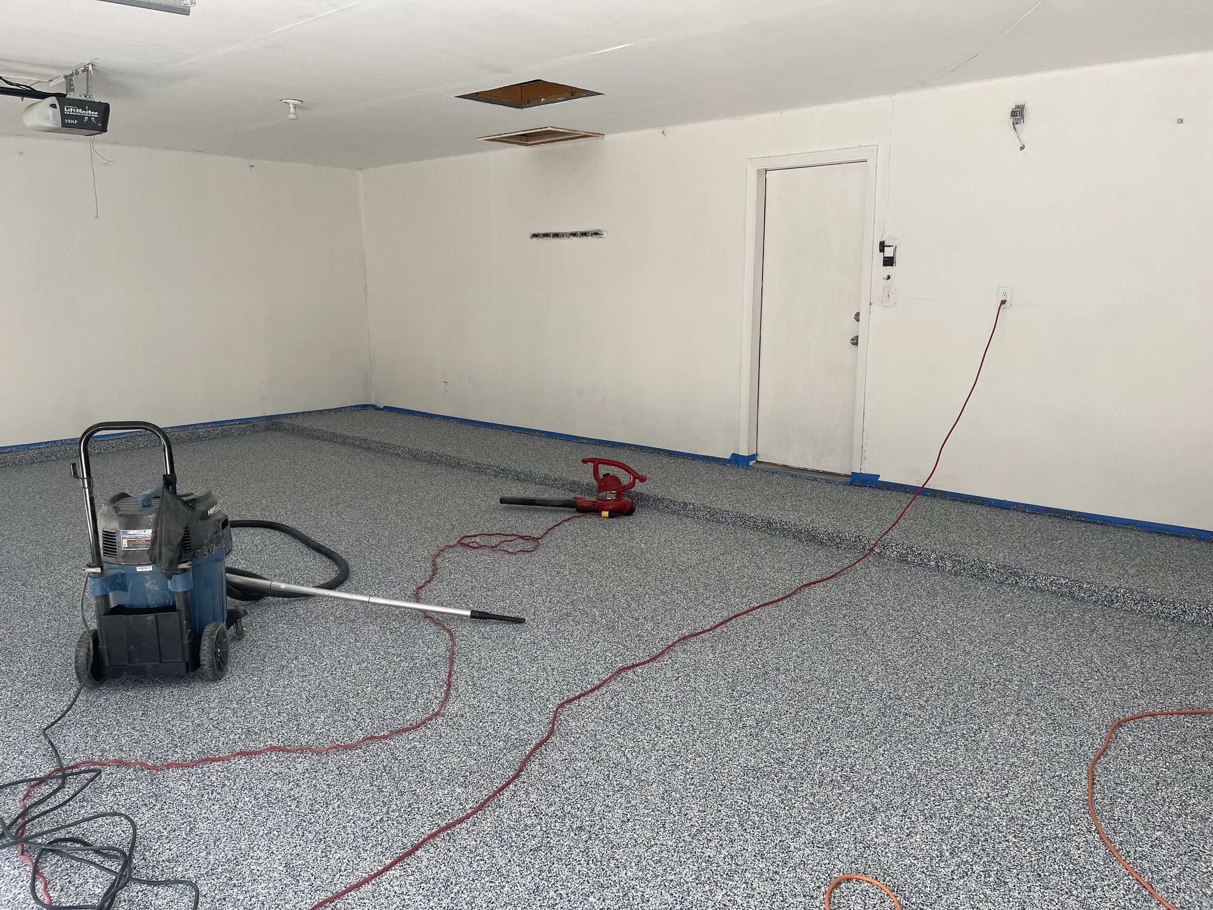 Empty garage with speckled gray flooring, a closed white door, and tools including a shop vacuum and a handheld sander on the floor. Blue painter's tape along the base of the walls and ceiling with exposed ceiling panels.