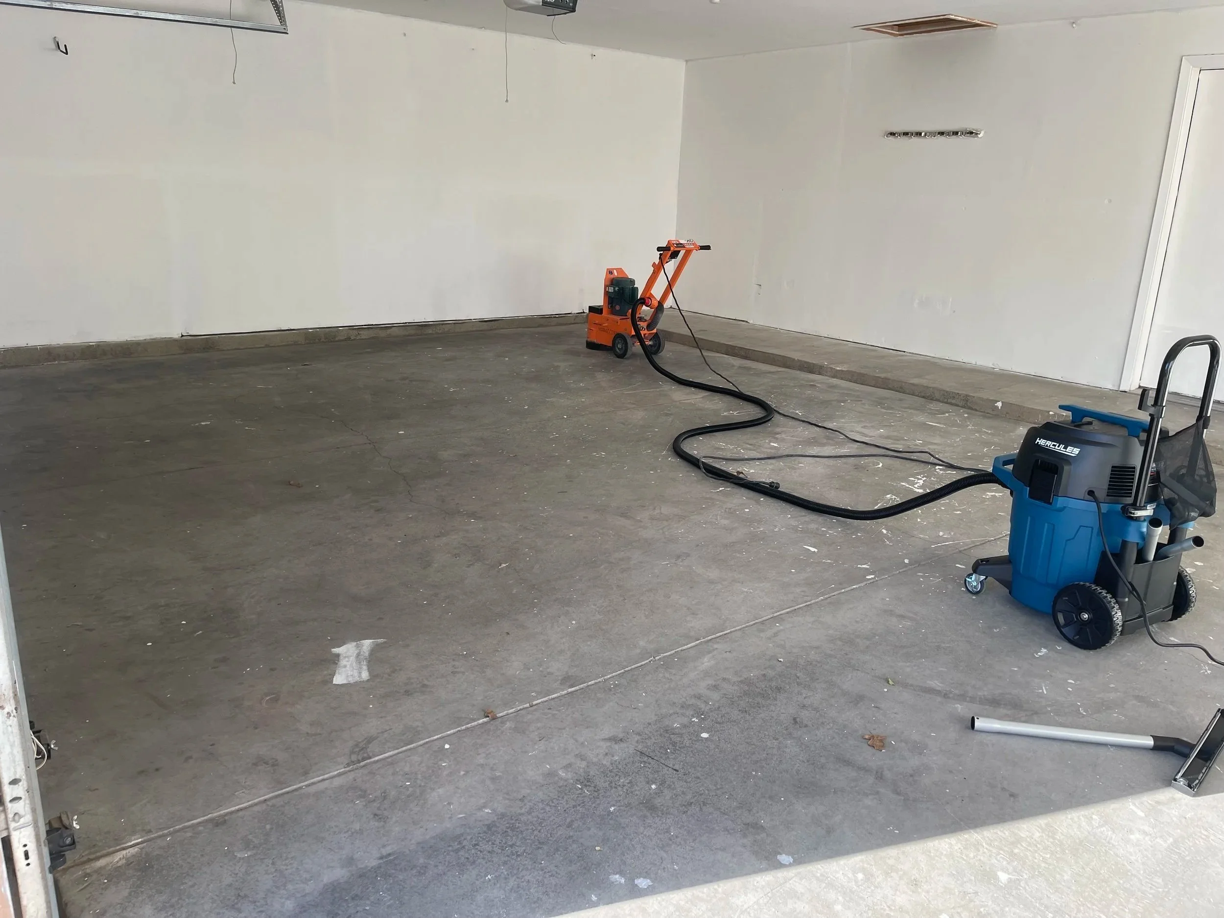 Empty garage with a concrete floor. There are two floor cleaning machines, one orange and one blue, connected by hoses. The garage appears to be under cleaning or renovation.