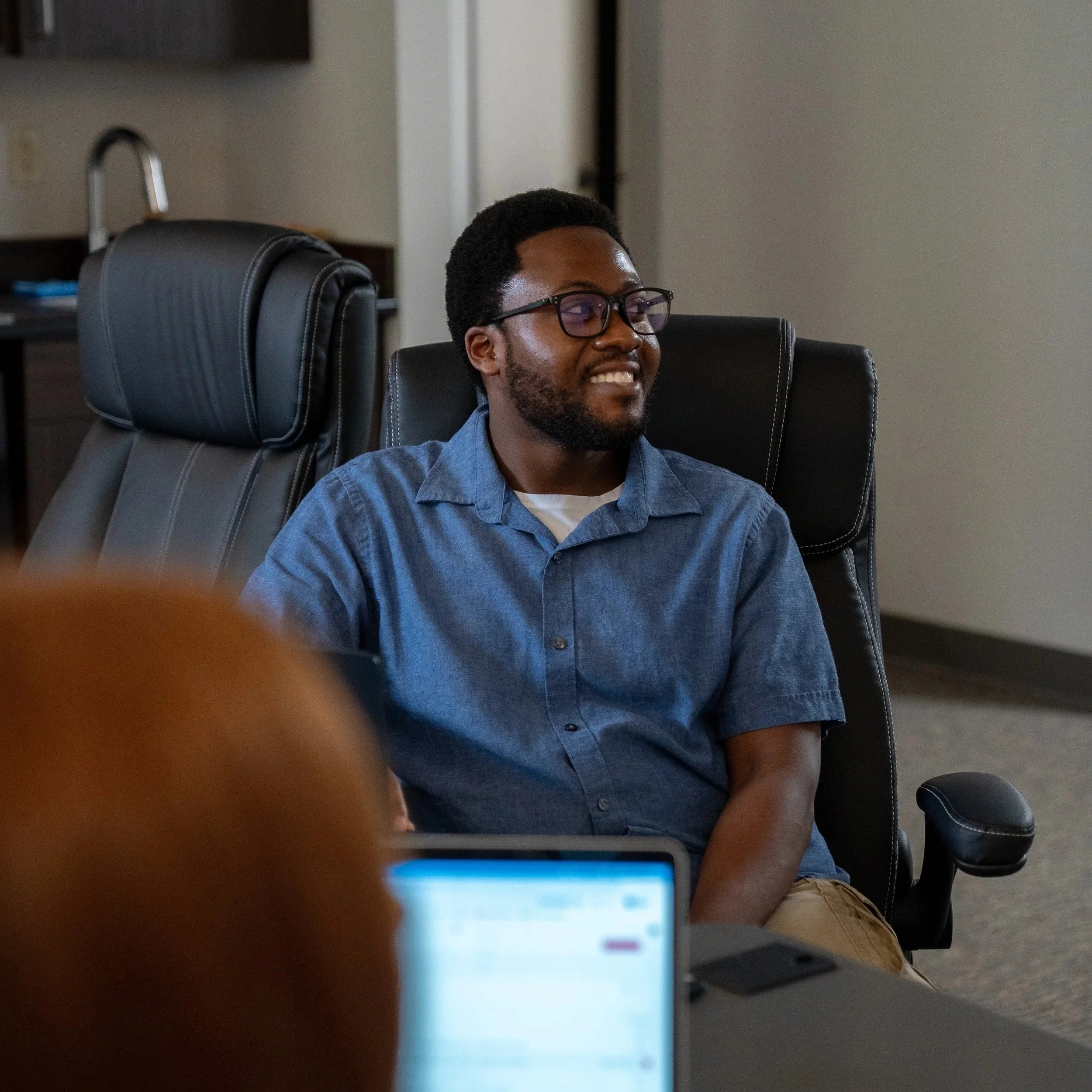 A person with glasses smiling, sitting in an office chair, with another person's finger partially visible in the foreground, and a computer screen in front.