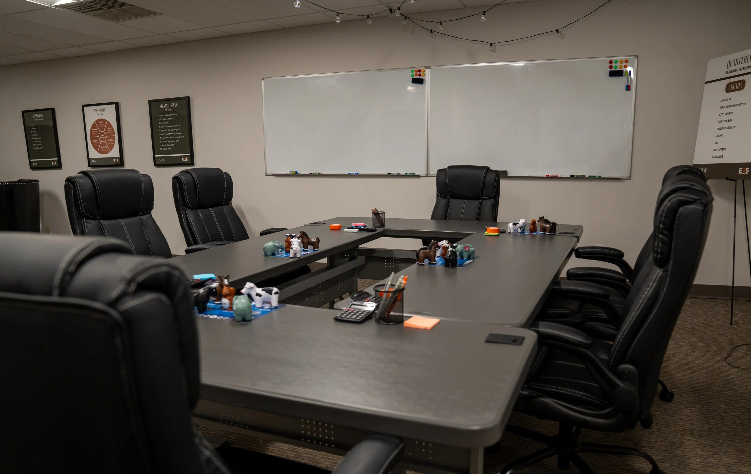 A conference room with tables and black leather chairs. The tables have small animal figurines, pens, and sticky notes. Three framed posters are on the wall, and a whiteboard is centered on the wall with markers on the tray. String lights hang from the ceiling.