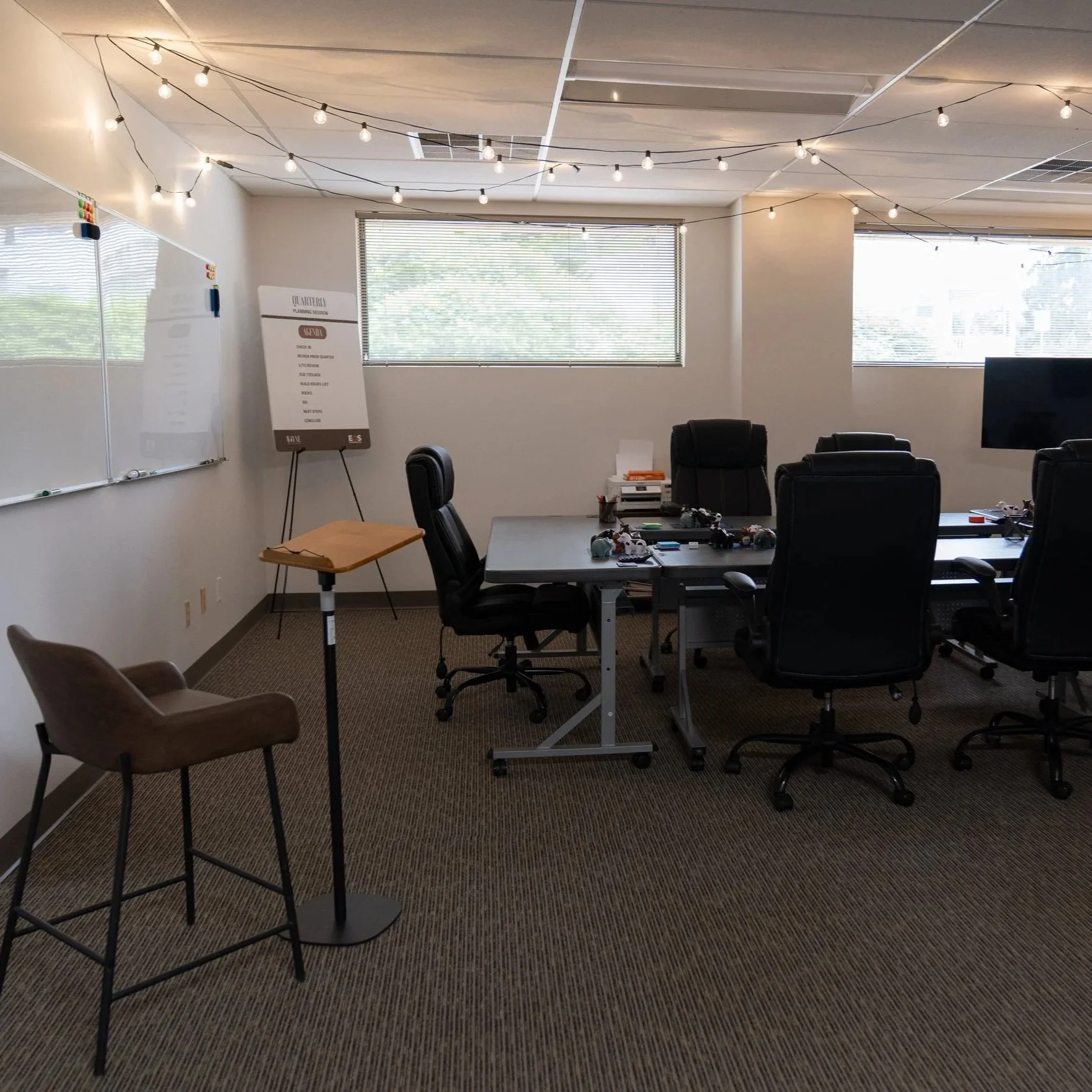 A modern conference room with a long table, multiple office chairs, a whiteboard on the wall, and string lights hanging from the ceiling. There are large windows with blinds allowing natural light. The room contains various items on the table, includ
