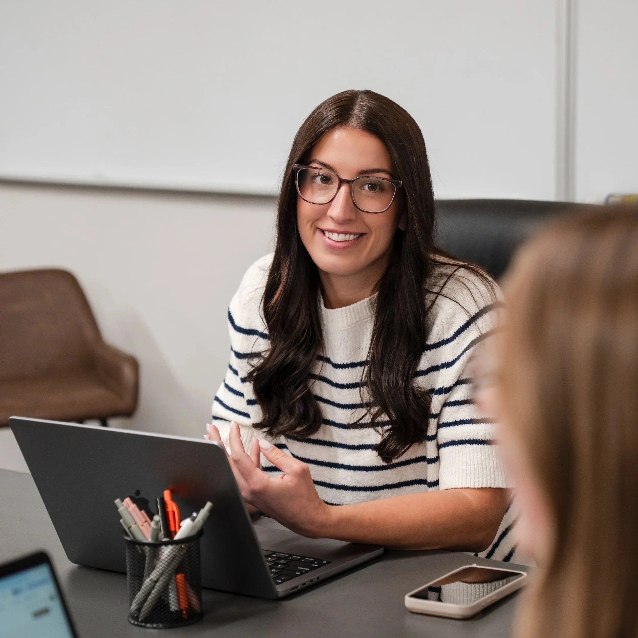 A woman with long dark hair and glasses smiling at her desk, working on a laptop surrounded by office supplies and electronic devices.