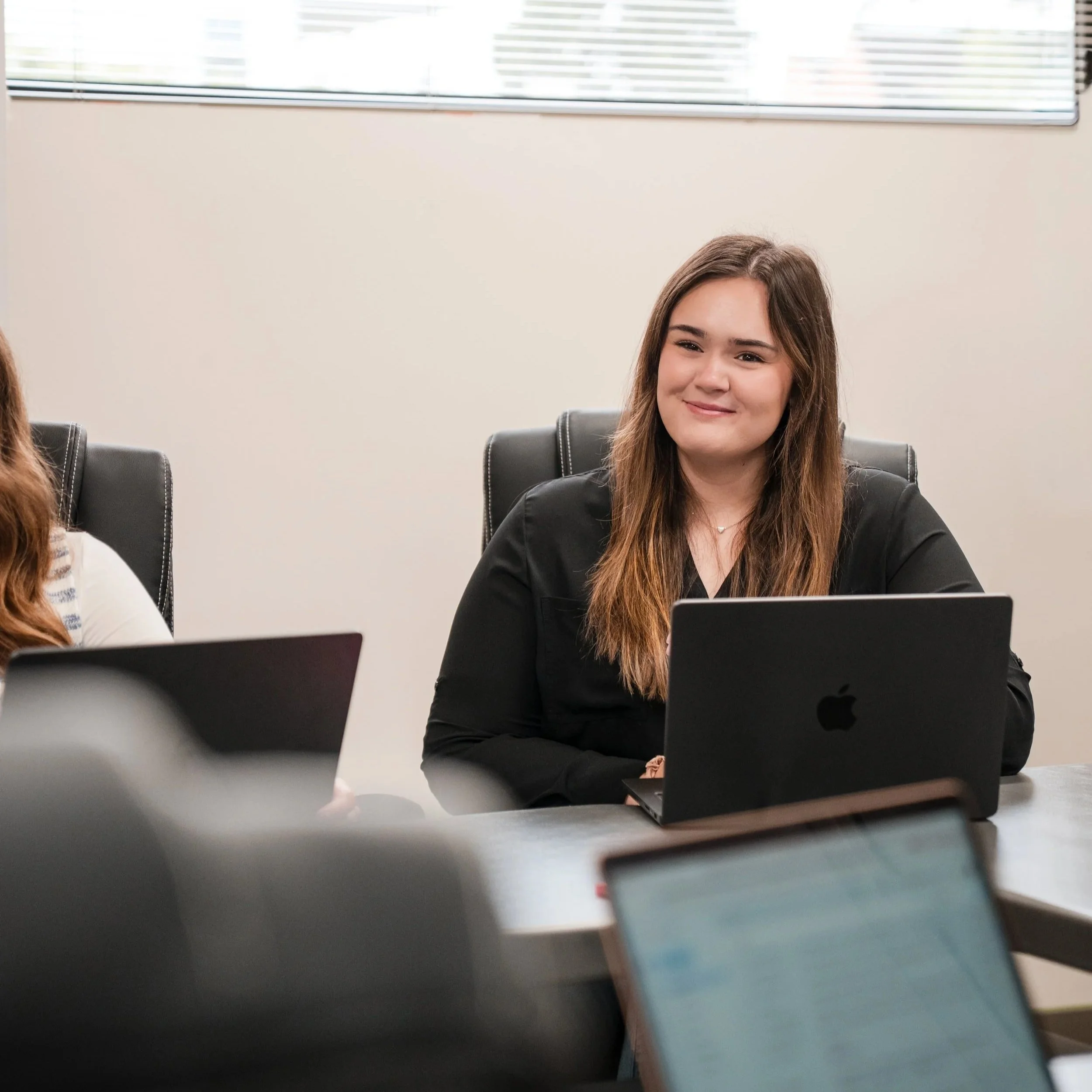 Young woman with long brown hair sitting in an office, smiling while looking at a computer.