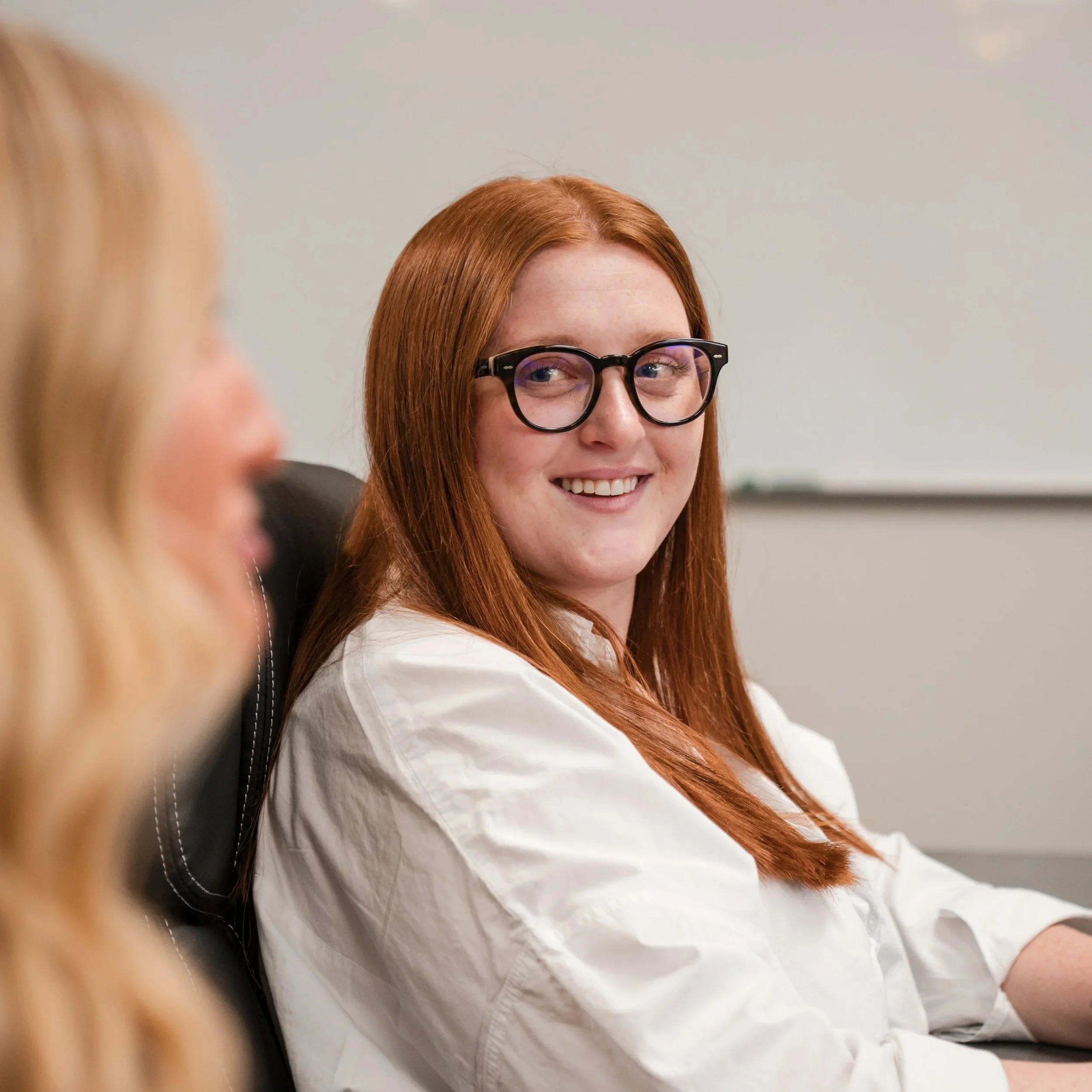 Close-up of a young woman with red hair, wearing glasses, smiling and looking slightly to her right, sitting in an indoor setting.