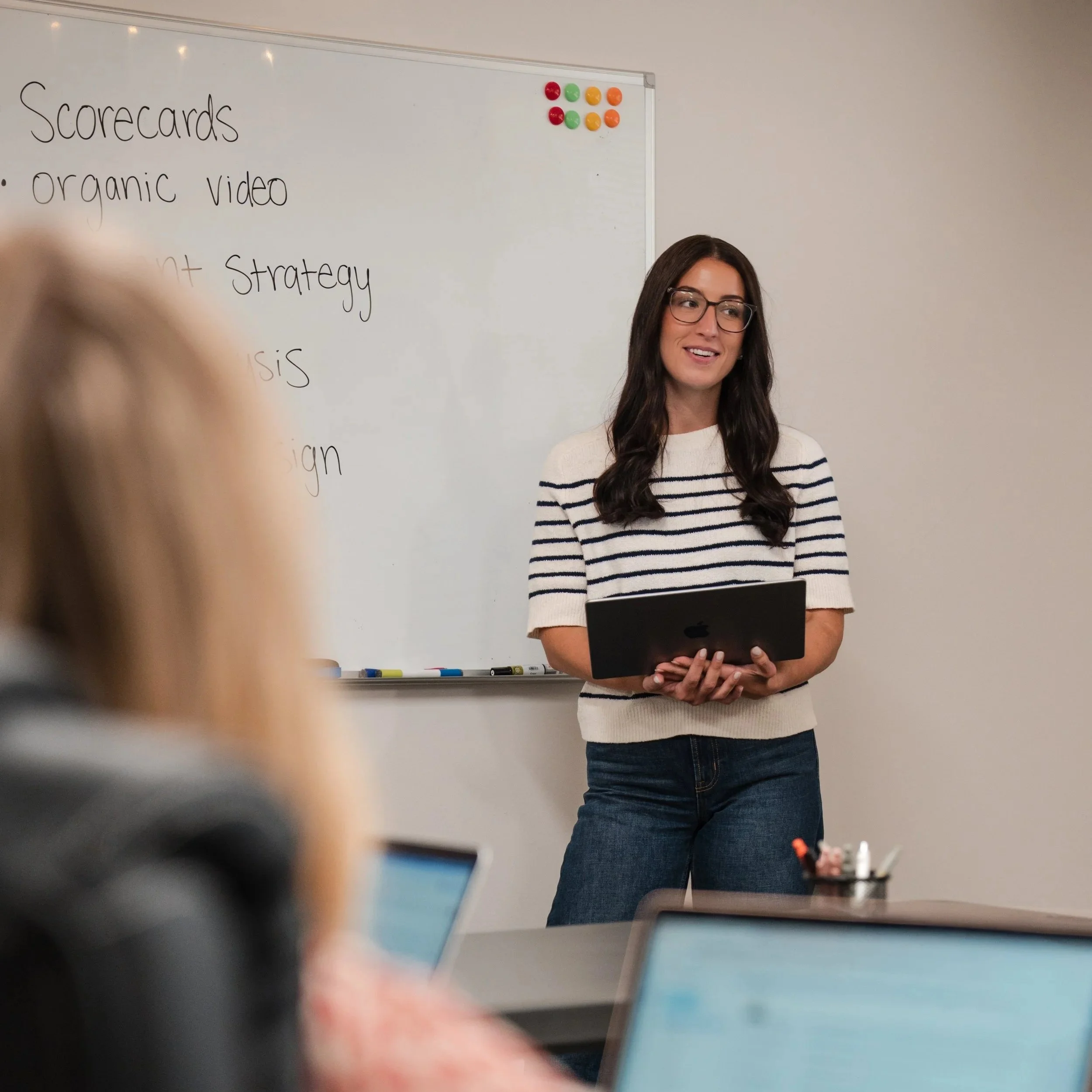 A woman with long dark hair, glasses, and a striped sweater, standing in front of a whiteboard with writing and colorful magnets, holding a laptop.