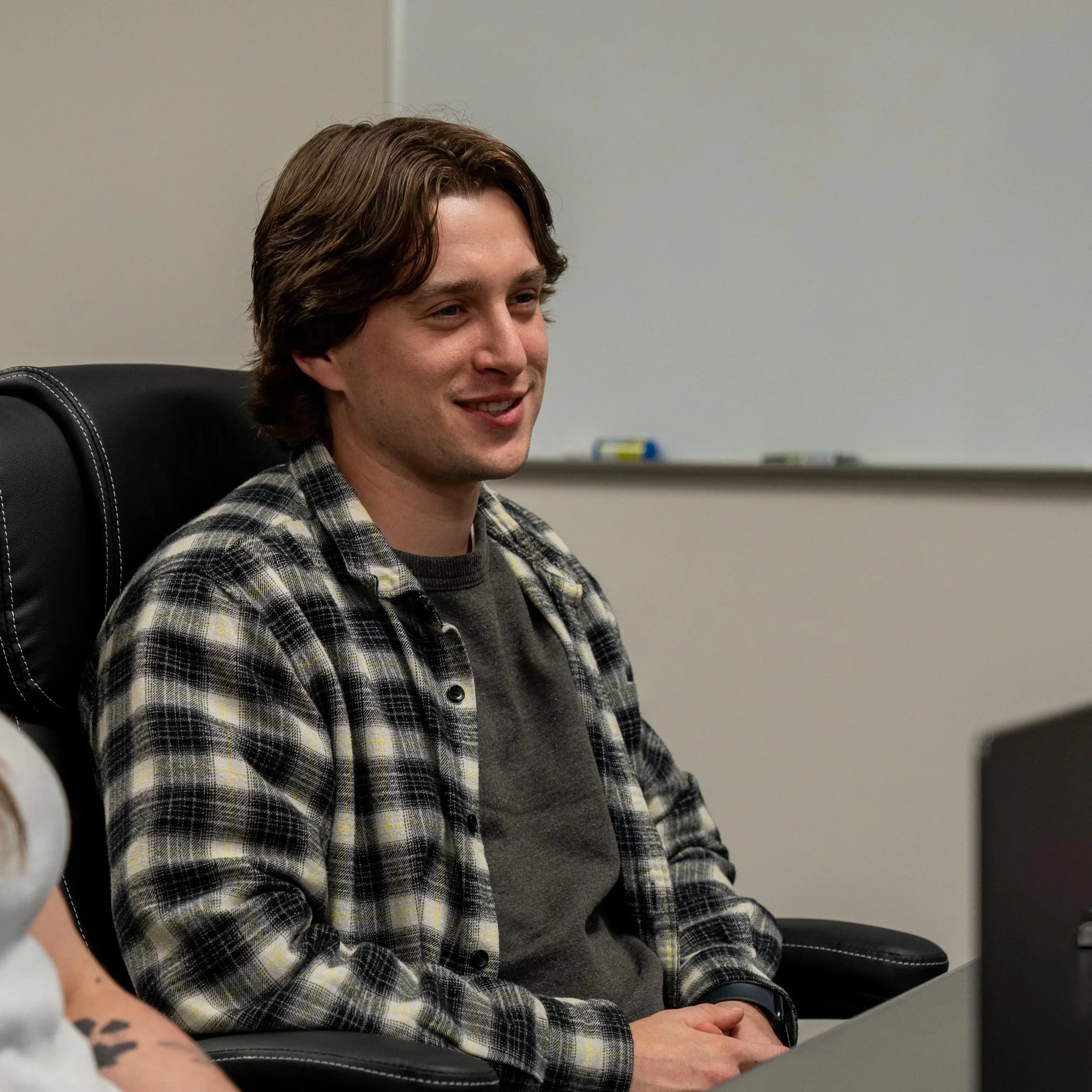 A young man with brown hair and a checked shirt sitting in a black leather chair, smiling, in an office setting.