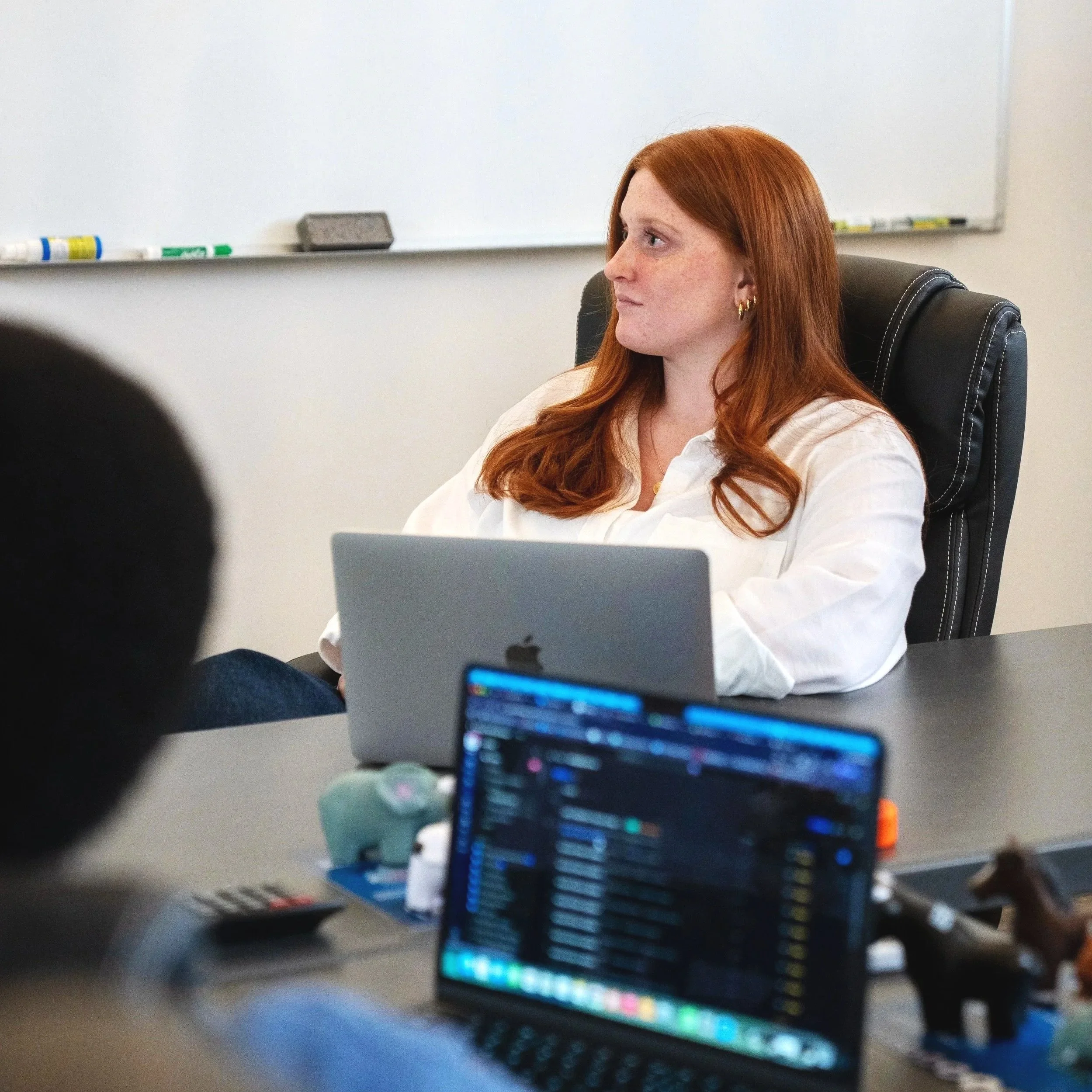 A woman with long red hair sitting at a desk in front of a whiteboard, looking to the side, with a laptop partially visible in the foreground.