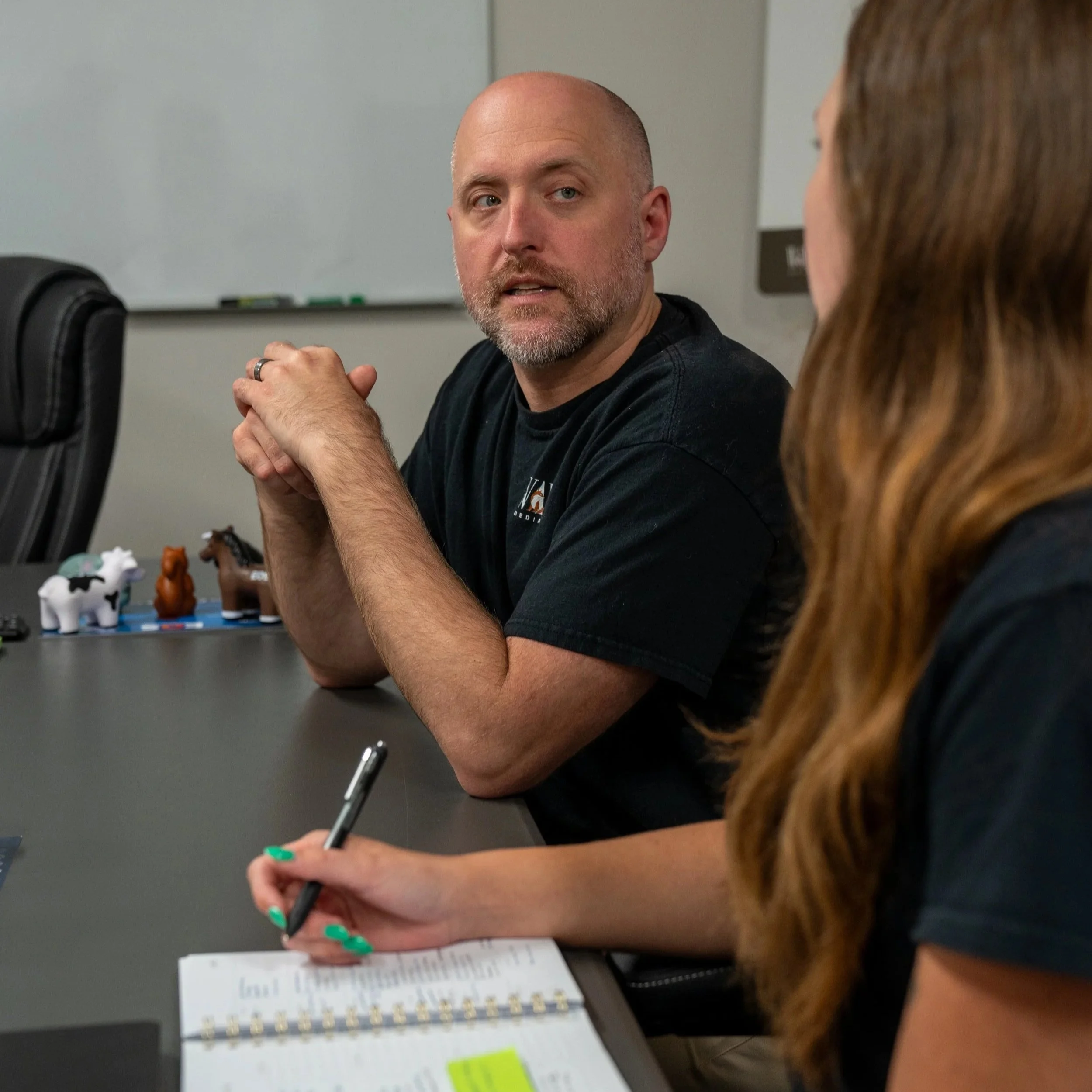 A man with a beard and a woman with red hair sitting at a table in a classroom or office setting engaged in conversation, with papers and a pen on the table.