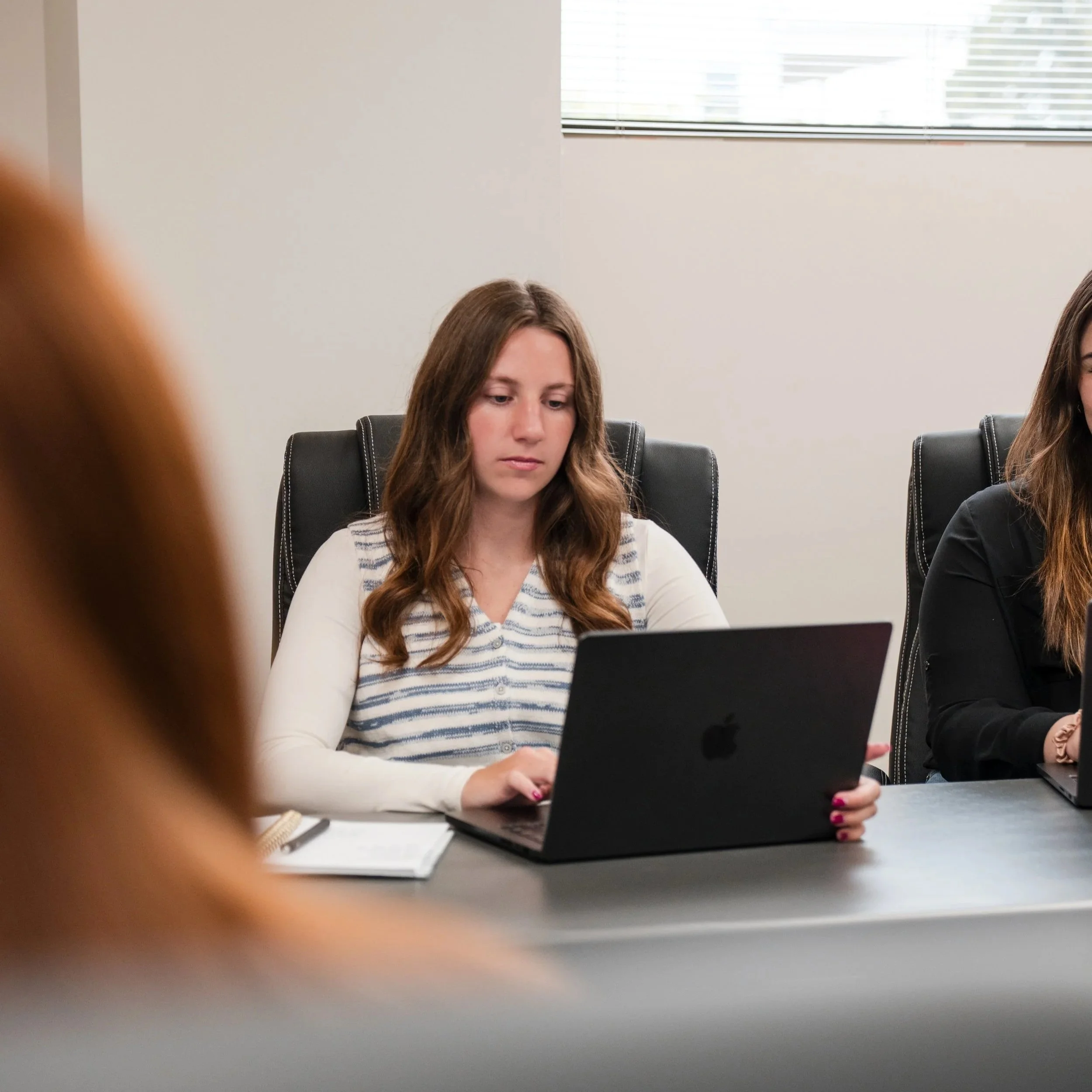 A young woman with brown hair sitting at a table, focused on a laptop. She is wearing a white and gray striped shirt. The background shows a white wall with a window with blinds, and part of another person is visible on the right side.