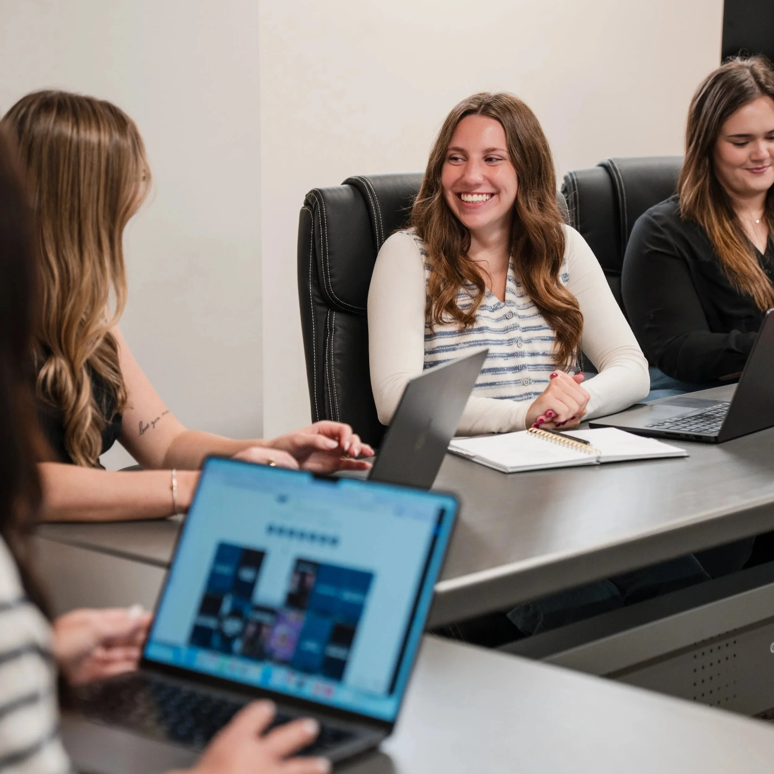 Multiple women sitting at a conference table, engaging in a discussion, some taking notes or using electronic devices, in an office setting.