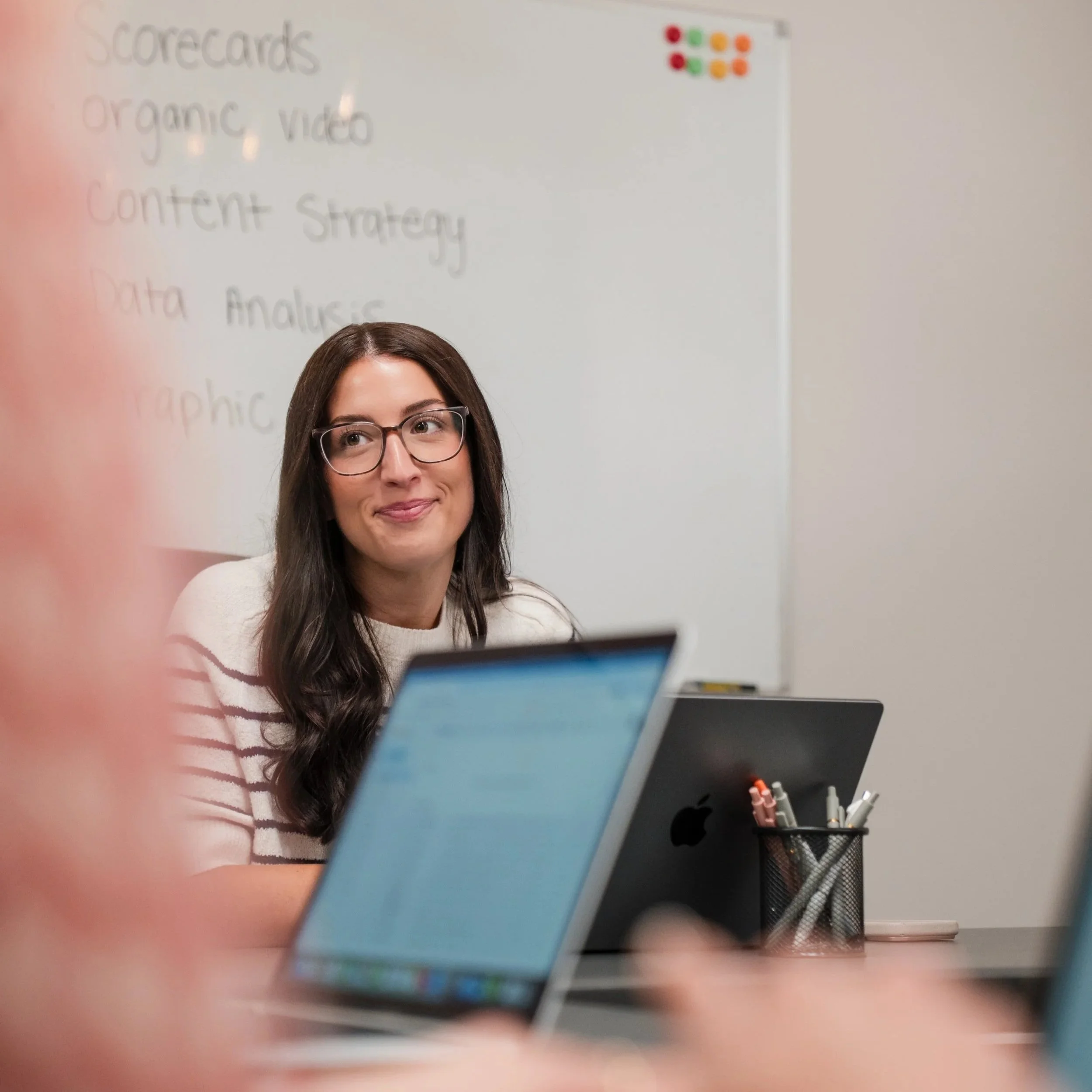 A young woman with long dark hair and glasses sitting at a desk with a laptop, looking upward. There is a whiteboard in the background with handwritten words related to content strategy, including 'scorecards', 'Organic Video', 'Content Strategy', 'D