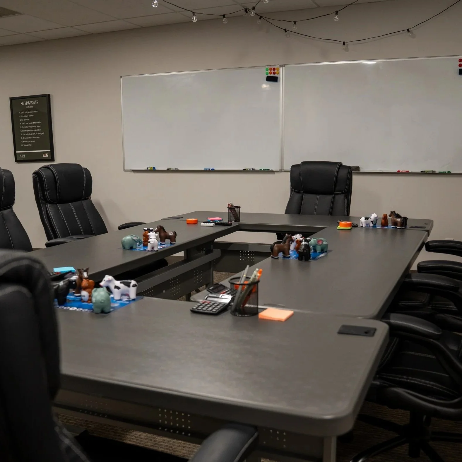 An empty conference room with black leather chairs around a long gray table, some small figurines of farm animals on blue mats, and a whiteboard in the background with a poster on the wall.