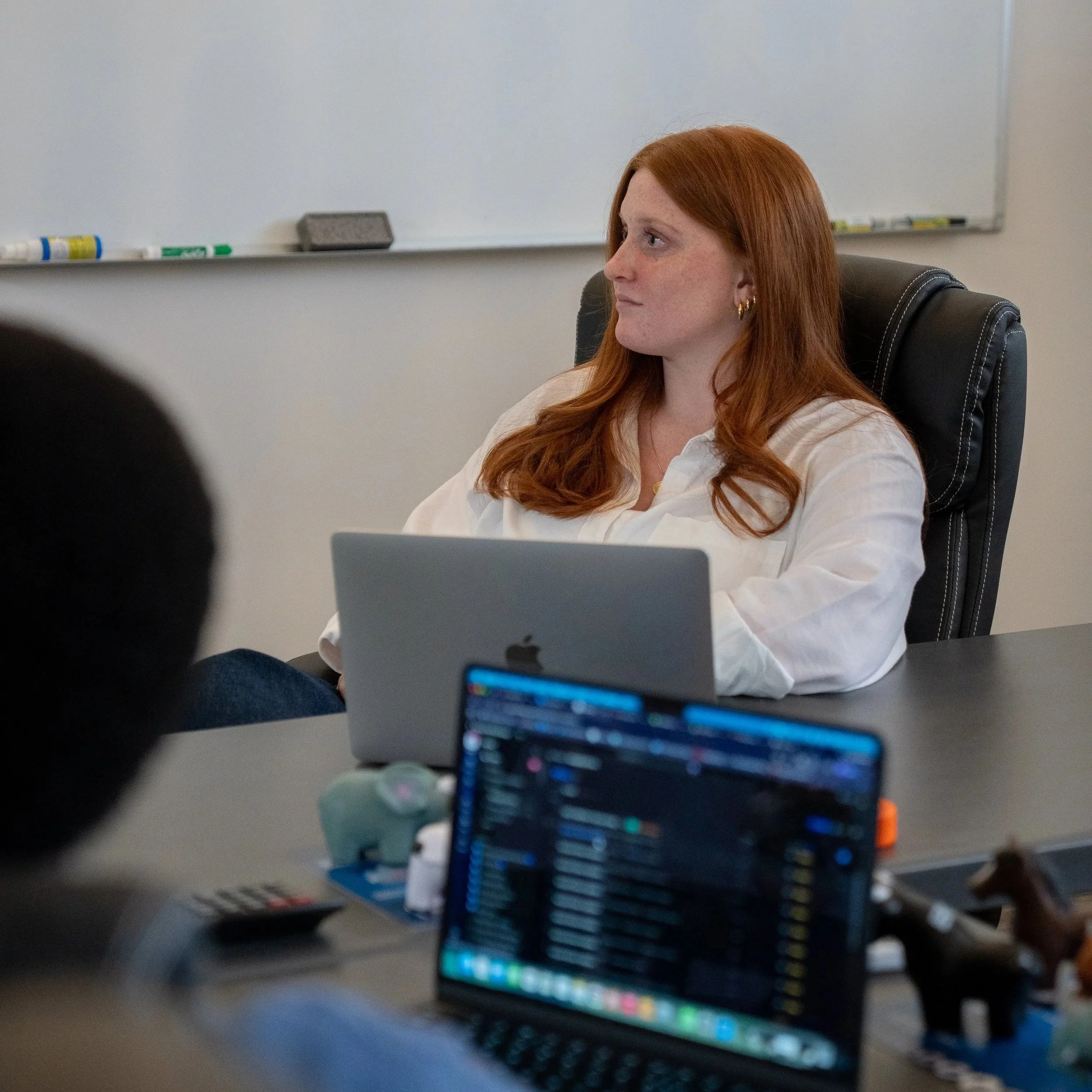 A woman with red hair sitting at a desk, with a whiteboard behind her, facing to the right. There are bottles of correction fluid on the whiteboard ledge and a laptop in front of her. The focus is on her profile.
